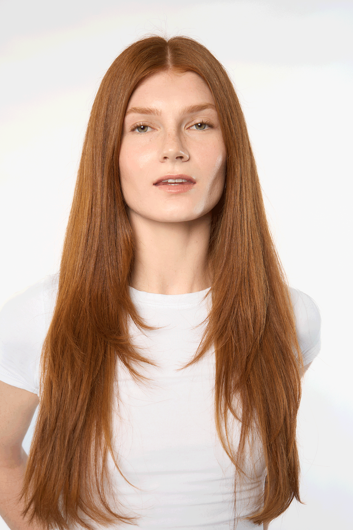 Young woman with long straight auburn hair wearing a white t-shirt poses against a light background.