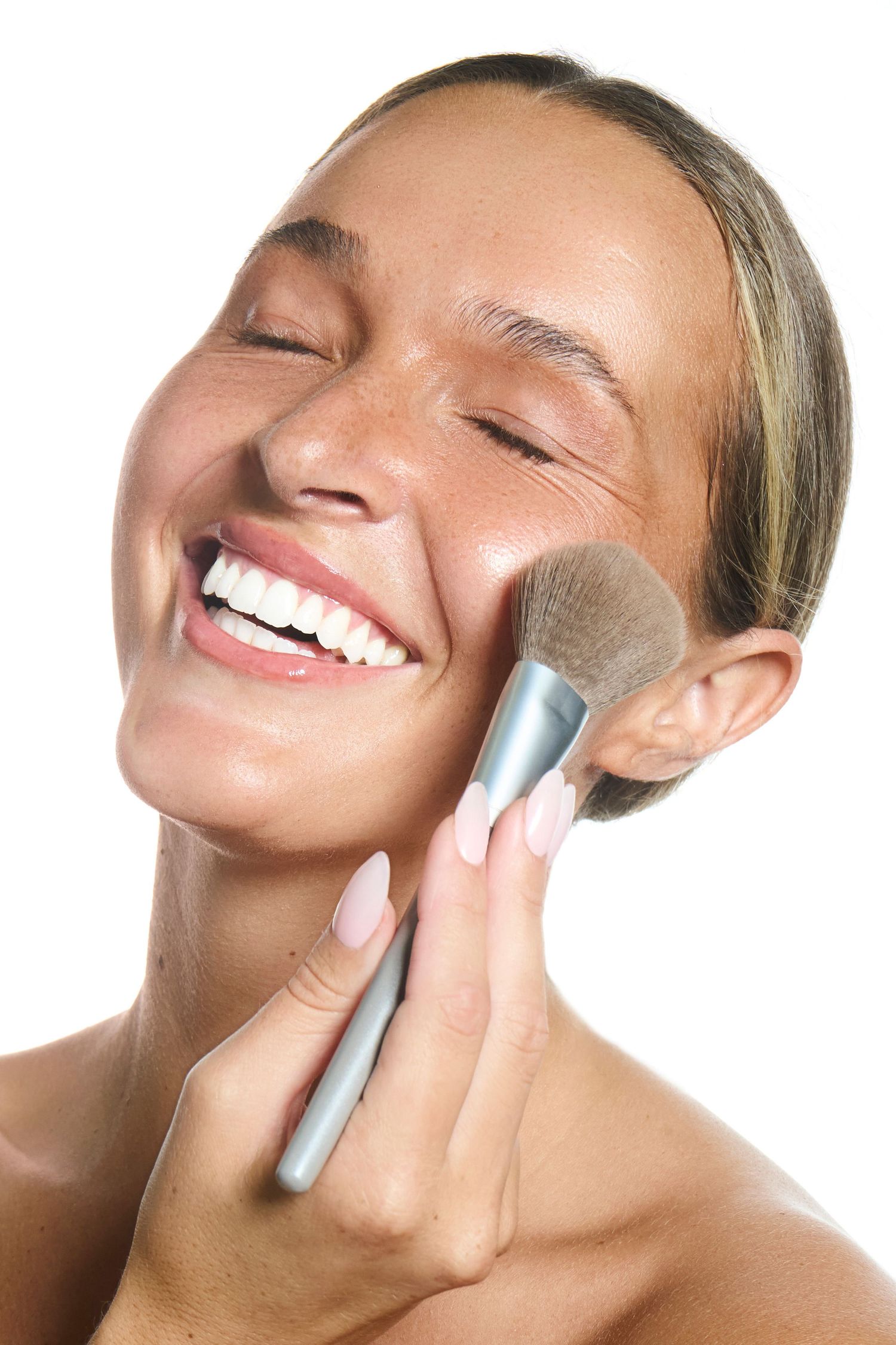 Smiling woman applying powder blush with a makeup brush on her cheek against a white background.