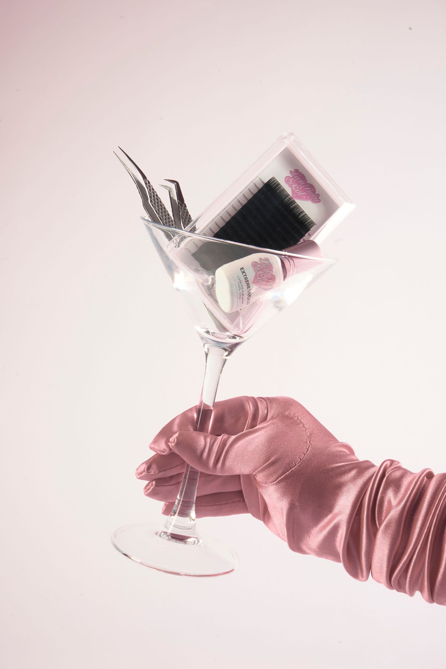 A gloved hand in pink satin holds a martini glass filled with beauty tools and eyelash accessories.