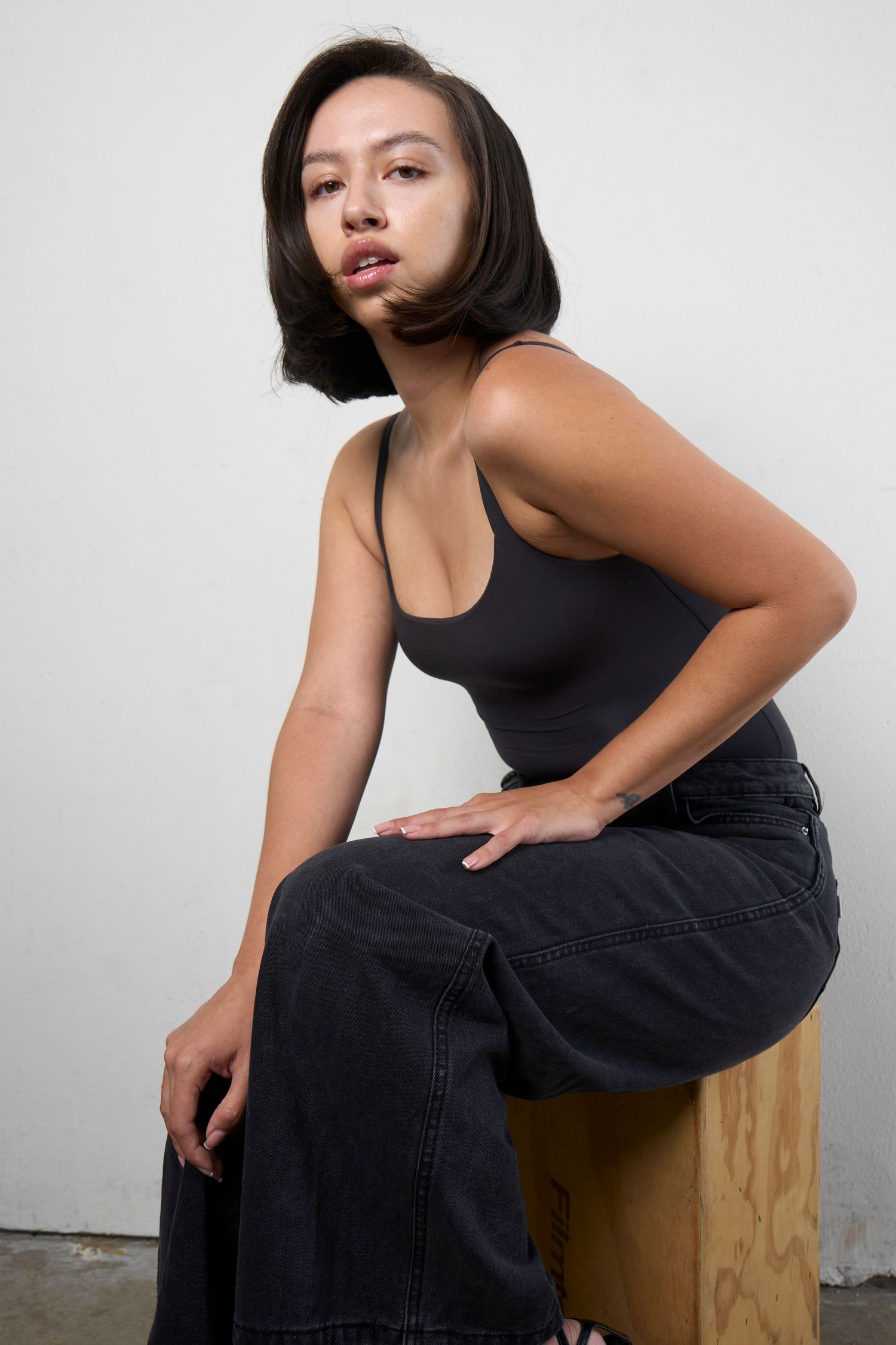 Young woman with bob haircut wearing black bodysuit and dark jeans, seated on wooden stool against white wall.