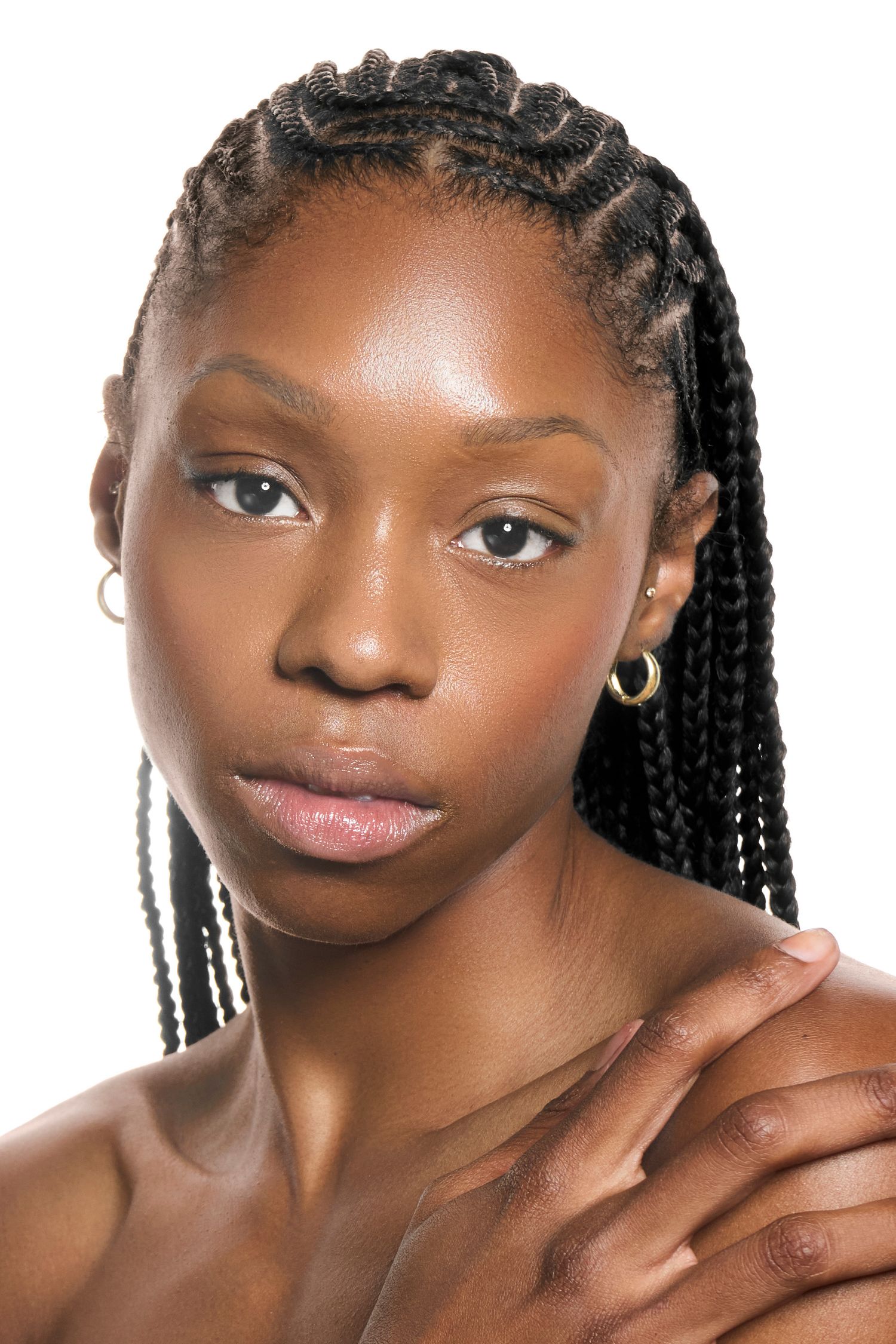 Young Black woman with box braids, gold hoop earrings, and glowing skin poses against white background.