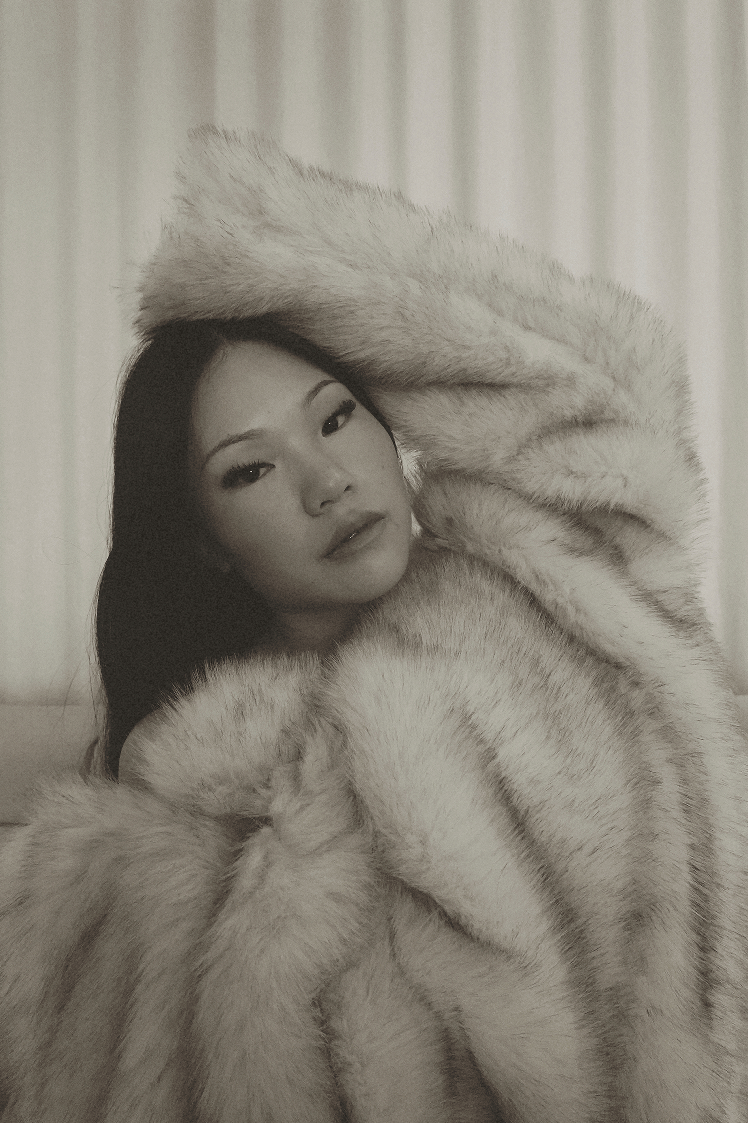 Black and white portrait of a young Asian woman draped in a luxurious fur coat, posing elegantly indoors.