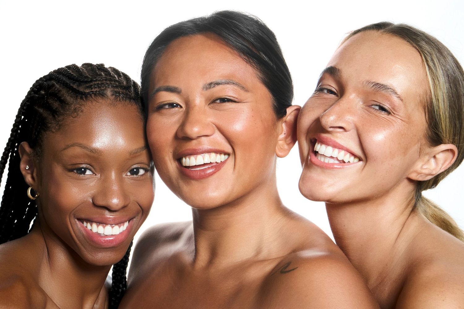 Three diverse smiling women with glowing skin posing together against a white background in a beauty portrait.