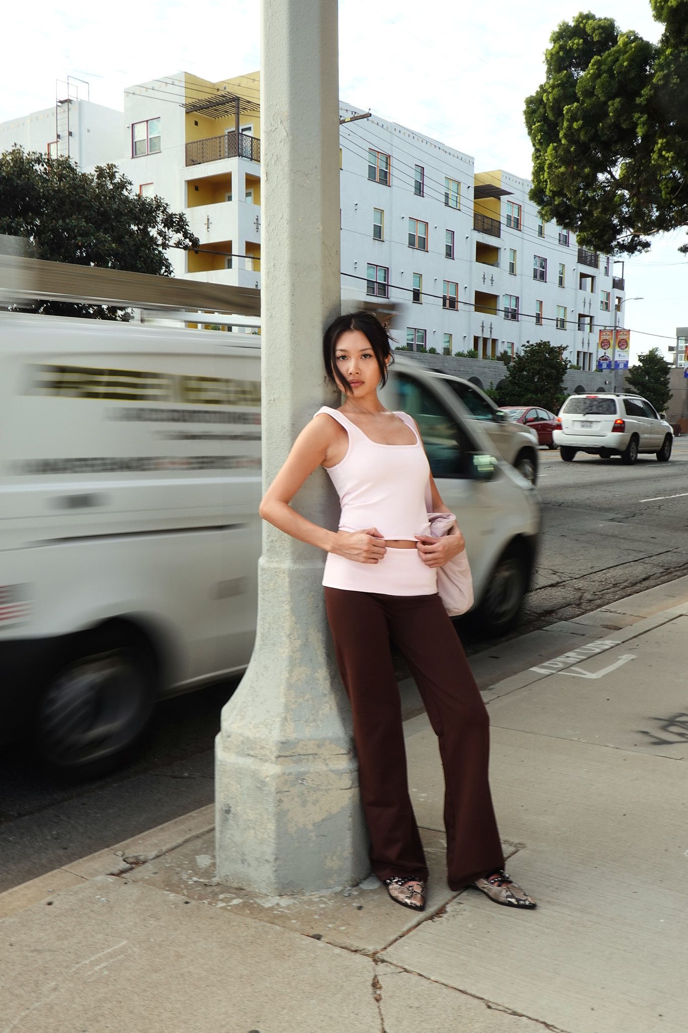 Young woman in pink tank top and brown flared pants leaning against a street pole on a busy urban sidewalk.