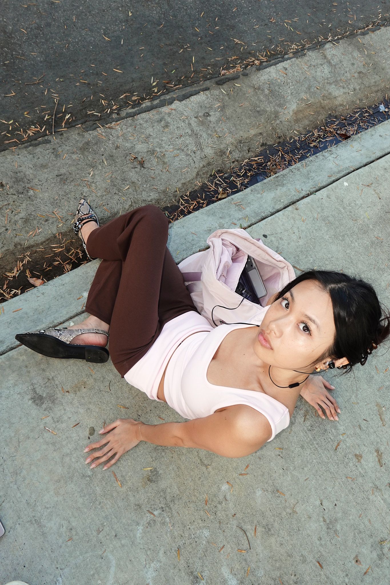 Young Asian woman in brown pants and white top sitting on a sidewalk curb, looking up at camera.