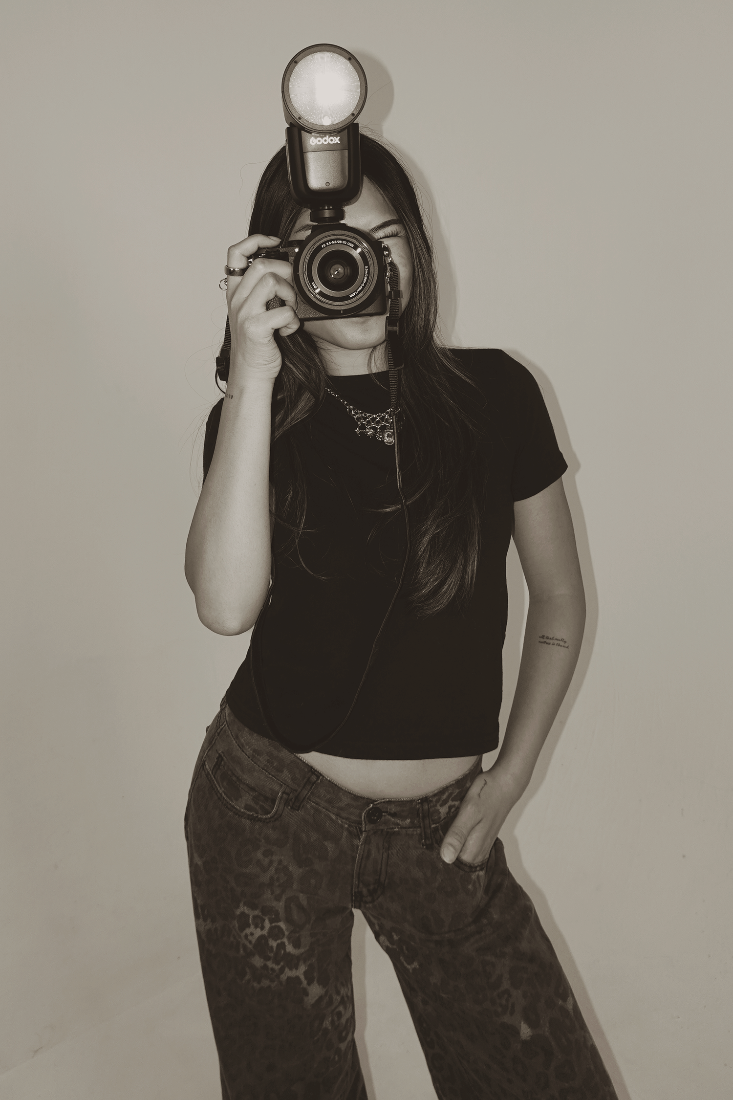 Sepia-toned photo of a young woman in black top and leopard jeans holding a DSLR camera with flash up to her face.