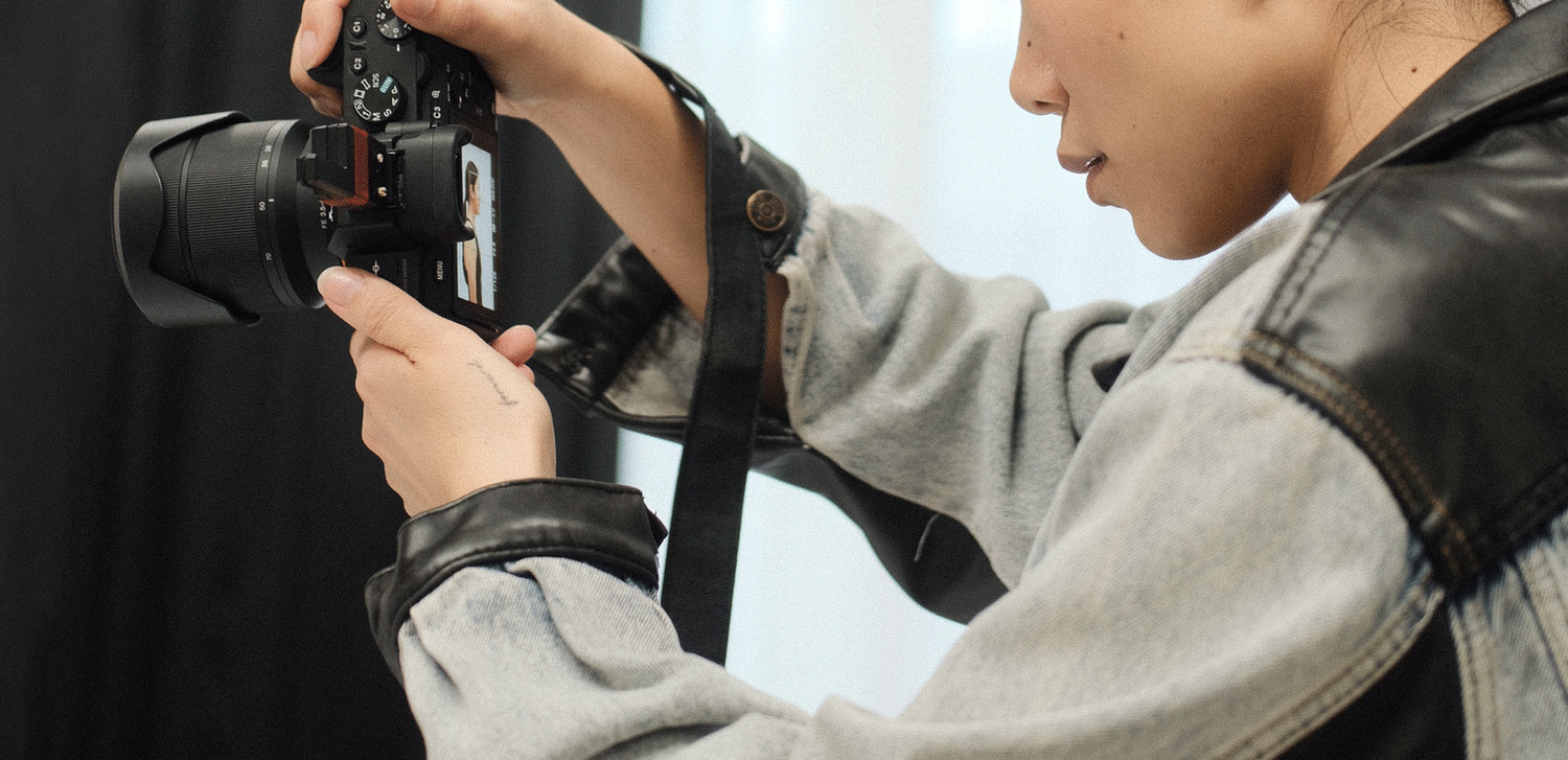 A young photographer examines their DSLR camera settings while wearing a denim jacket indoors.