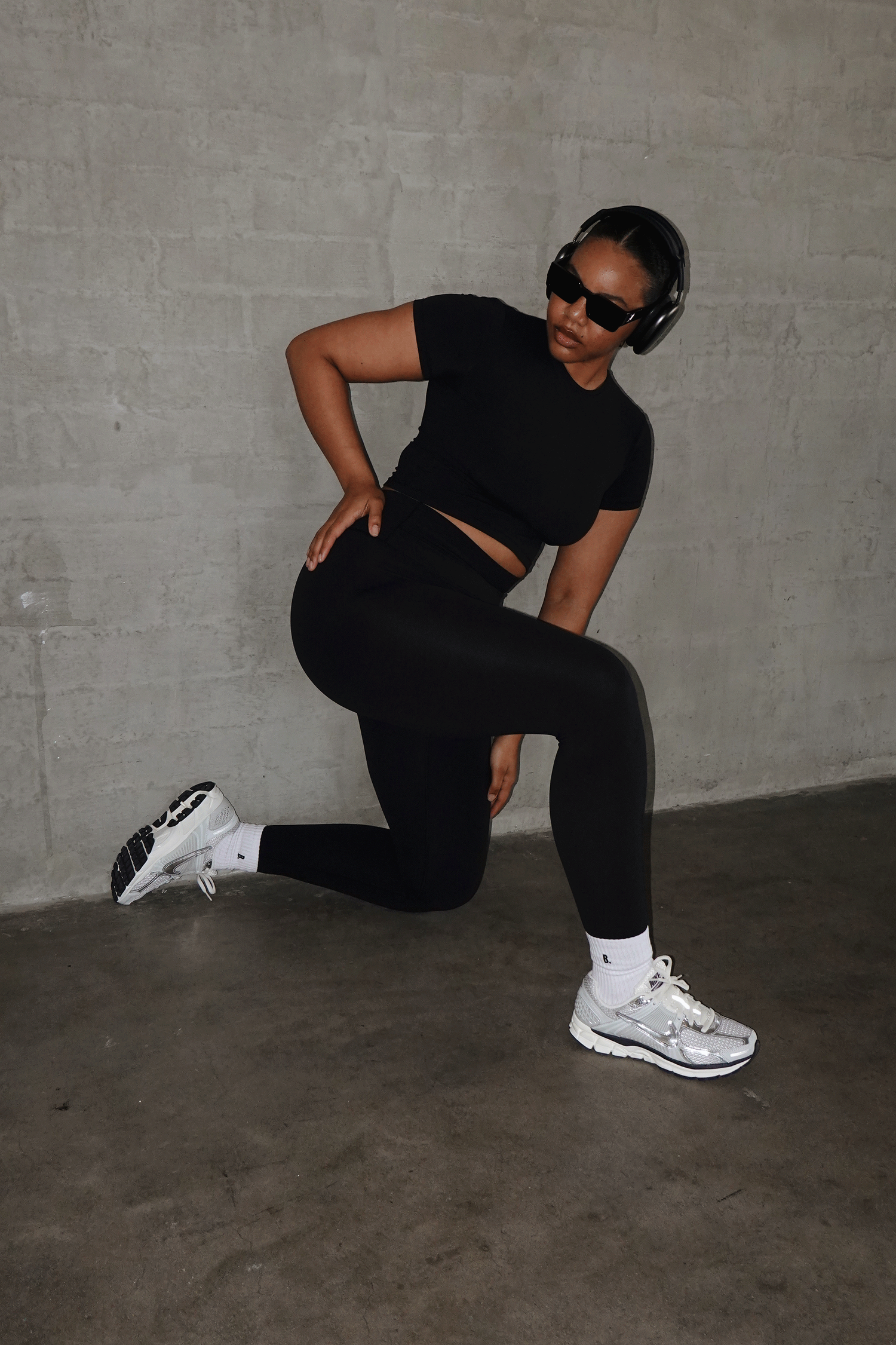 Woman in black crop top and leggings doing a lunge pose, wearing white sneakers, sunglasses, and headphones.