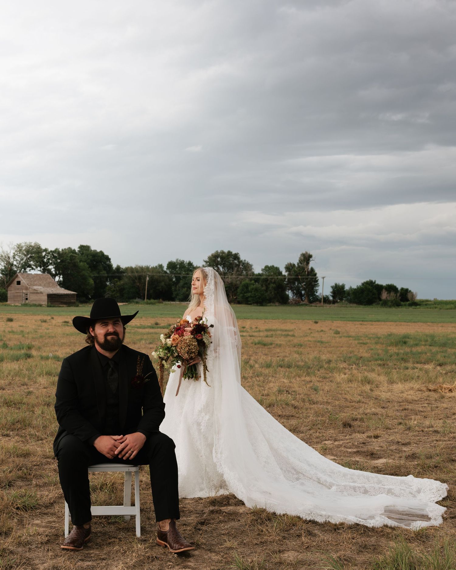Bride in lace gown with veil stands behind groom in black cowboy hat seated in field at rustic outdoor wedding.