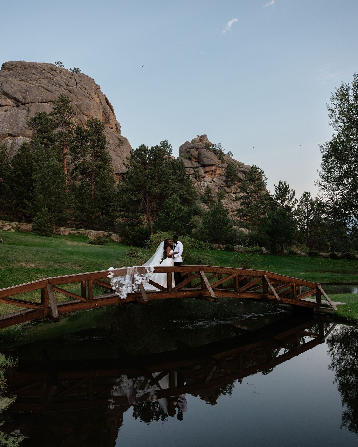 Bride and groom share romantic moment on wooden bridge reflected in pond with rocky mountain backdrop at dusk.