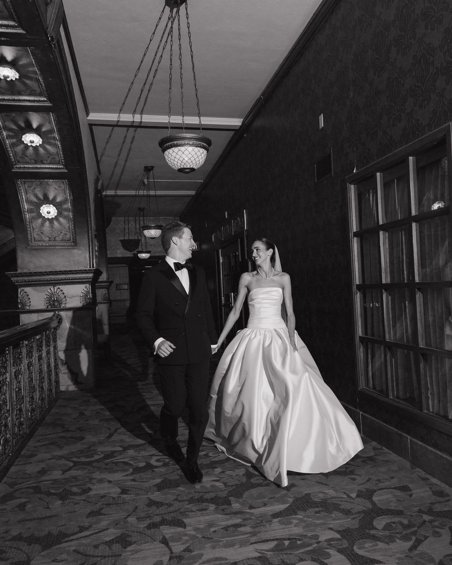 Bride and groom walking hand in hand through an ornate hotel corridor in black and white.