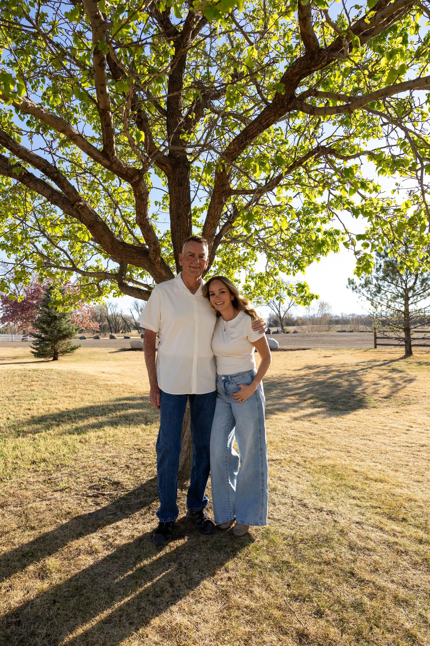 Couple posing together under a large tree in a park, man in white shirt and jeans with woman in light blue maxi skirt.