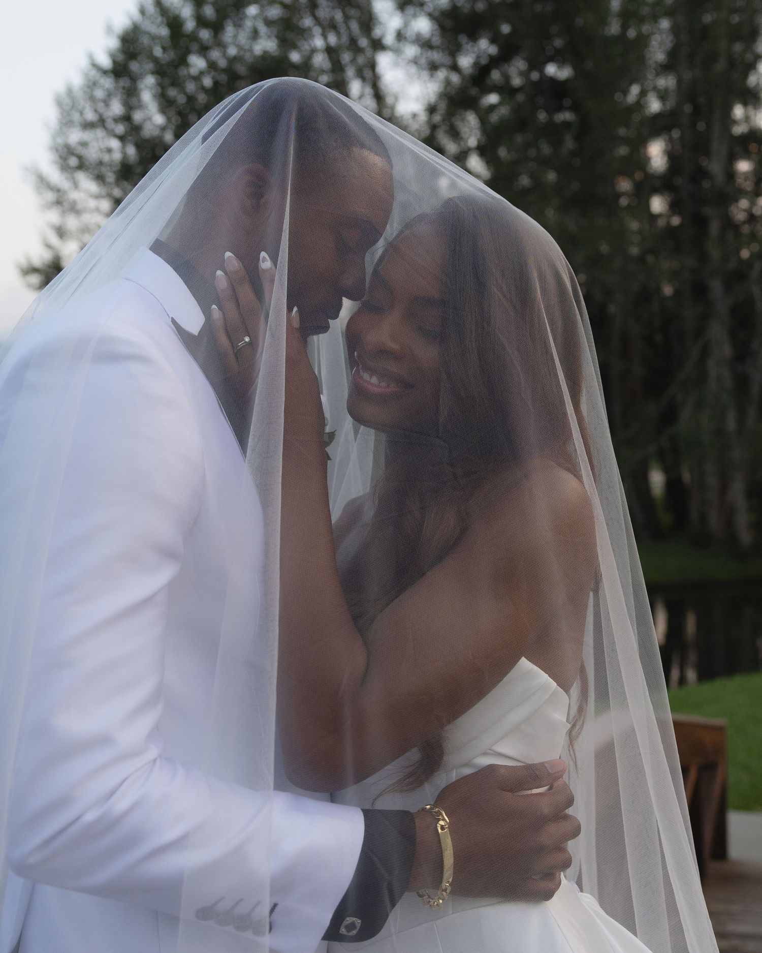 Newlyweds share intimate moment under bridal veil, smiling and embracing during outdoor wedding ceremony.
