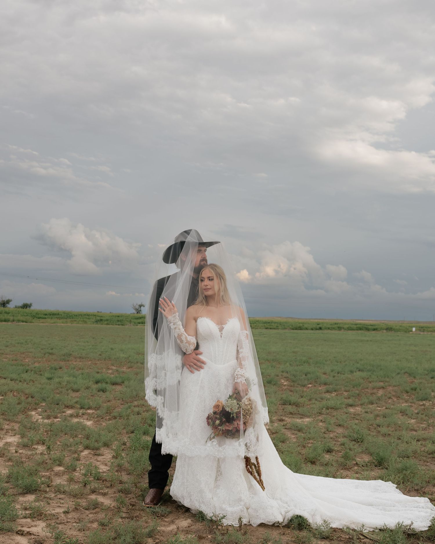 Bride in lace gown and groom in cowboy hat sharing intimate moment under veil in open field.