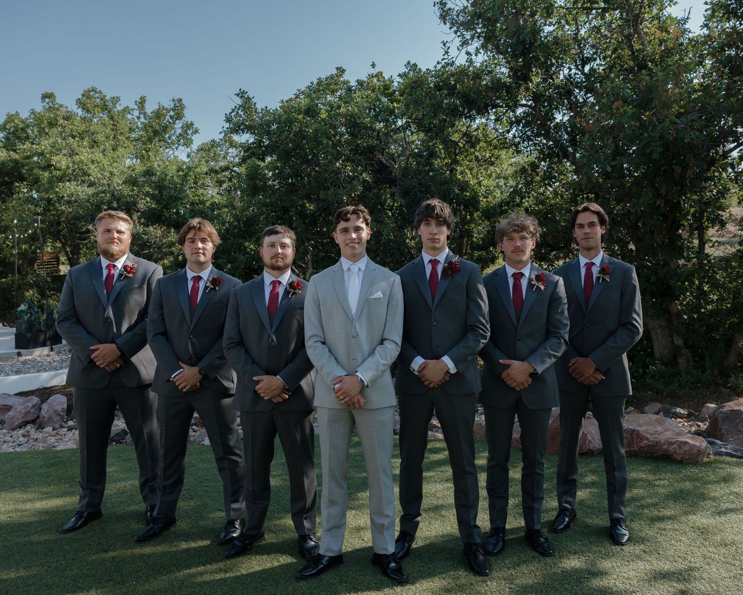 Groom in light gray suit stands with six groomsmen in dark suits and burgundy ties outdoors.