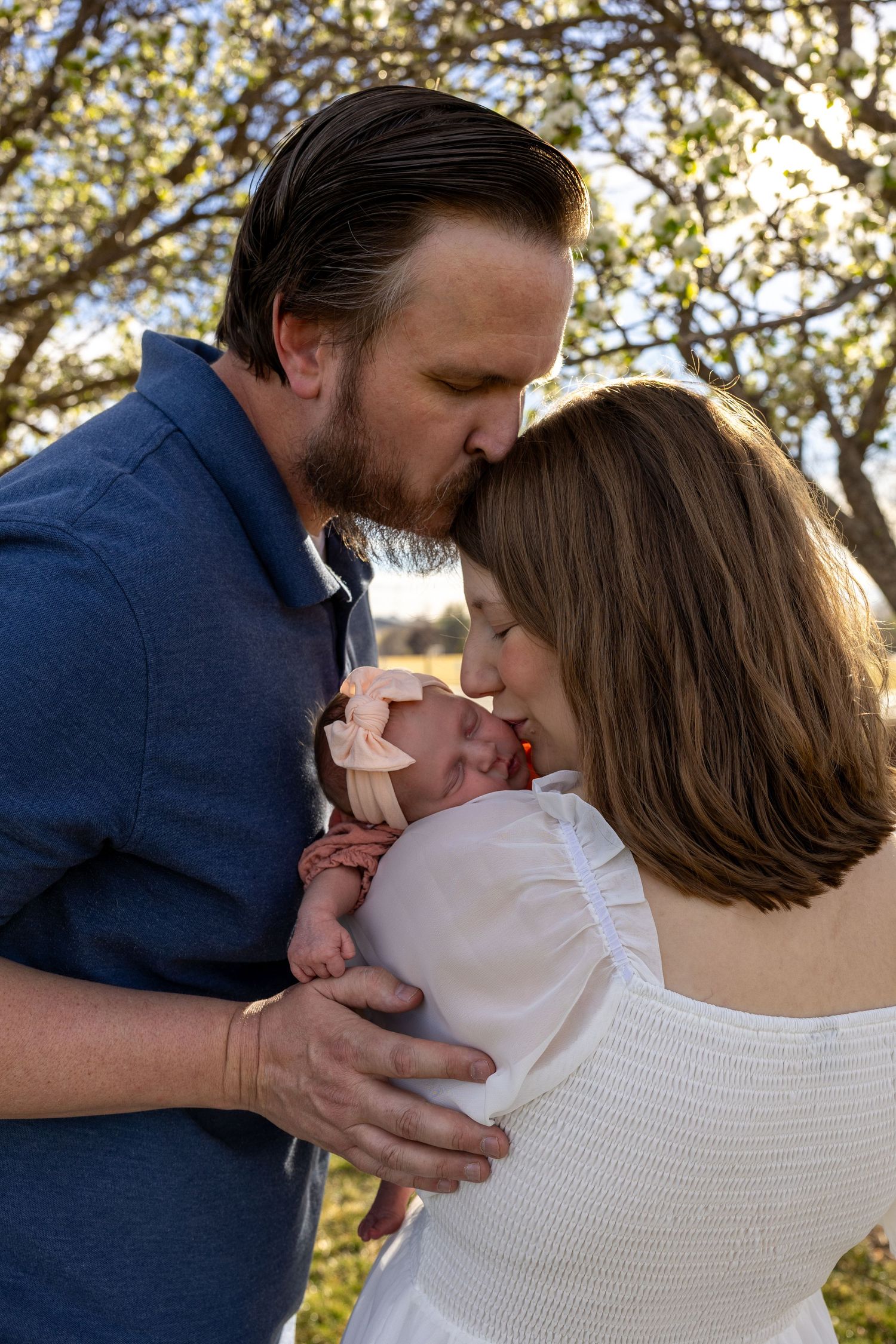 New parents tenderly kiss their newborn baby outdoors under blooming trees in warm golden sunlight. Family photography