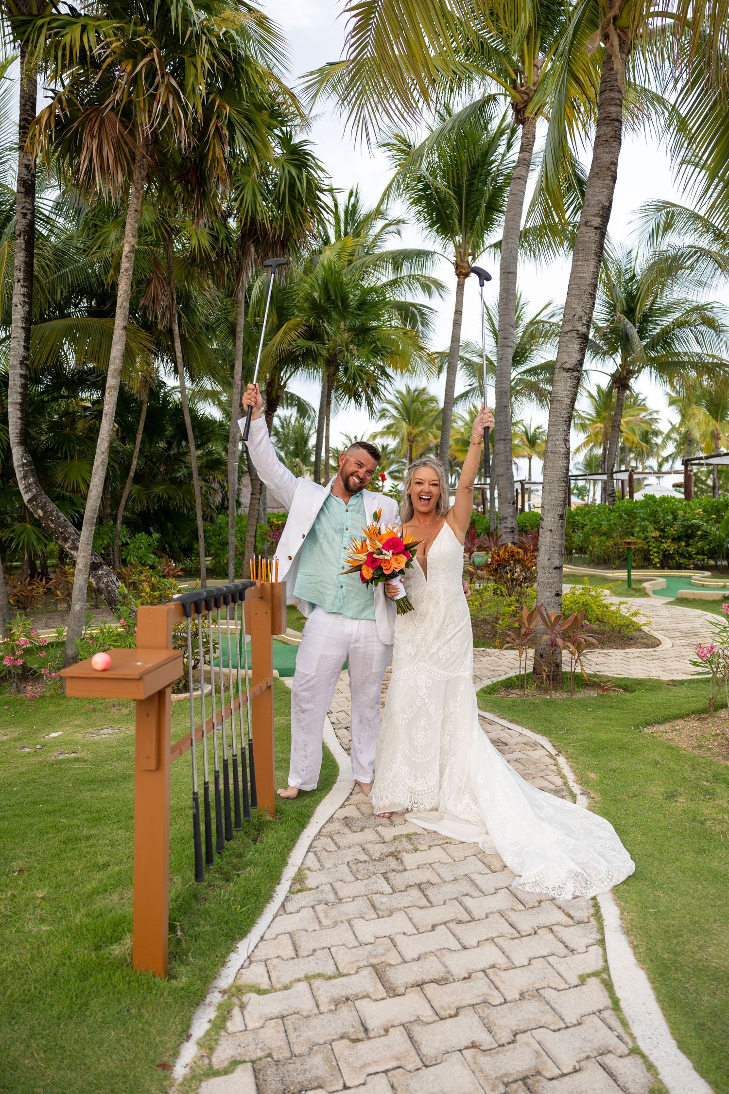 Joyful bride and groom celebrating on garden pathway holding golf clubs among lush tropical palm trees.