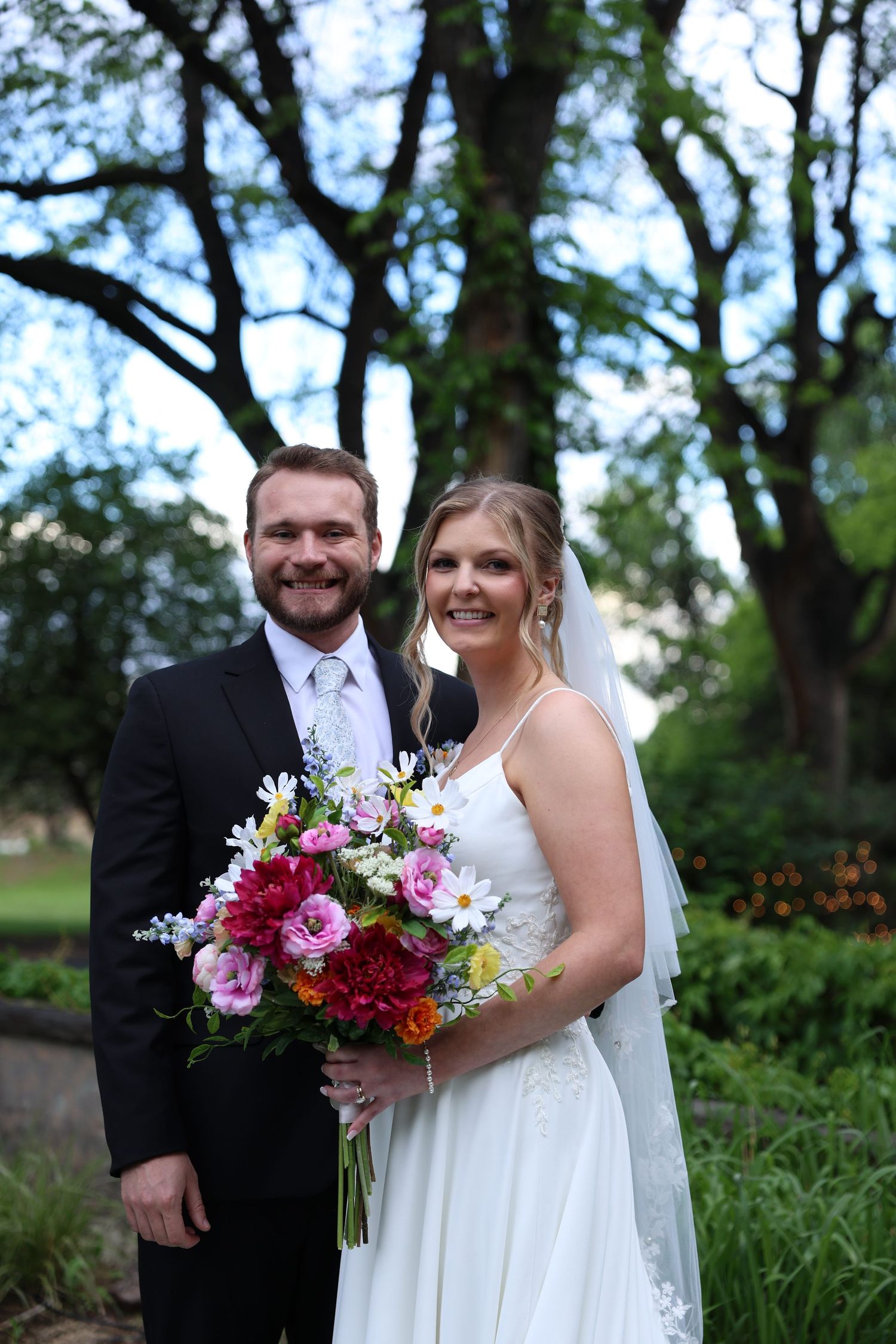Smiling bride and groom pose outdoors holding colorful wedding bouquet with pink and white flowers.