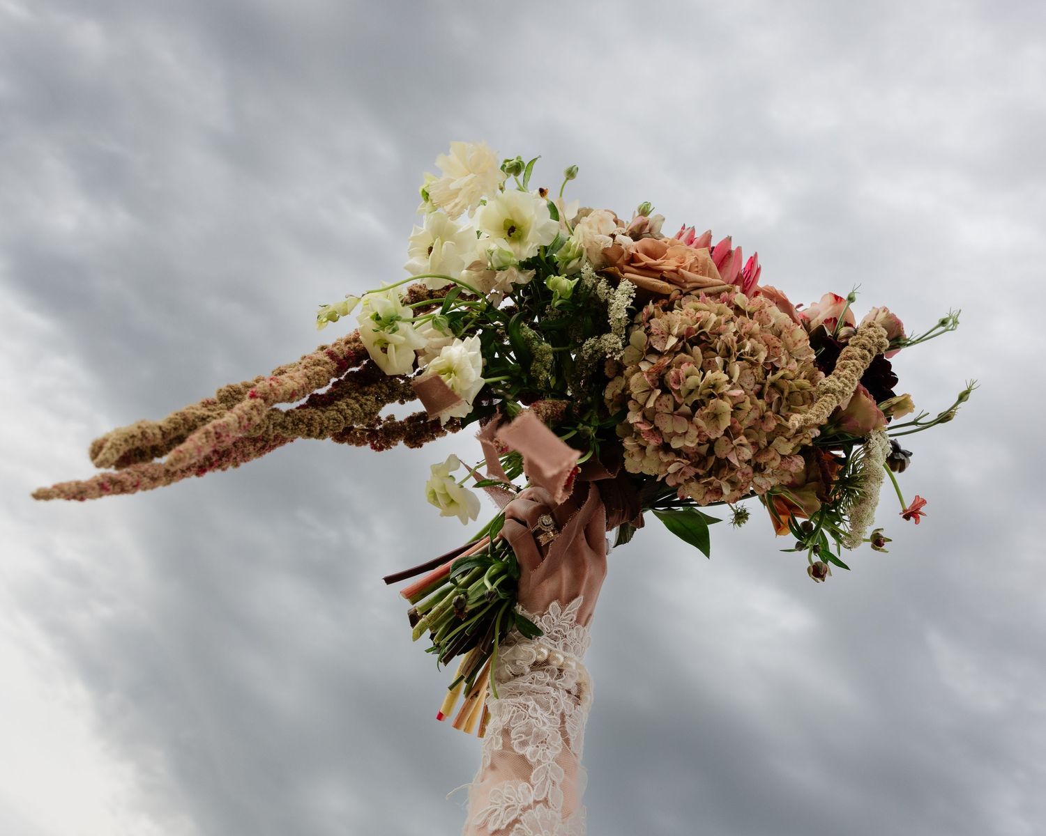 Rustic bridal bouquet with dried hydrangeas, amaranth, and white ranunculus held high against stormy sky.