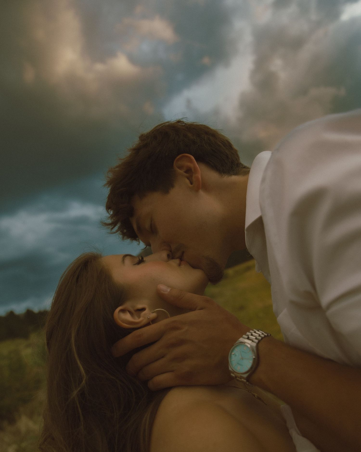 Romantic couple sharing a kiss under dramatic stormy clouds in an outdoor setting.