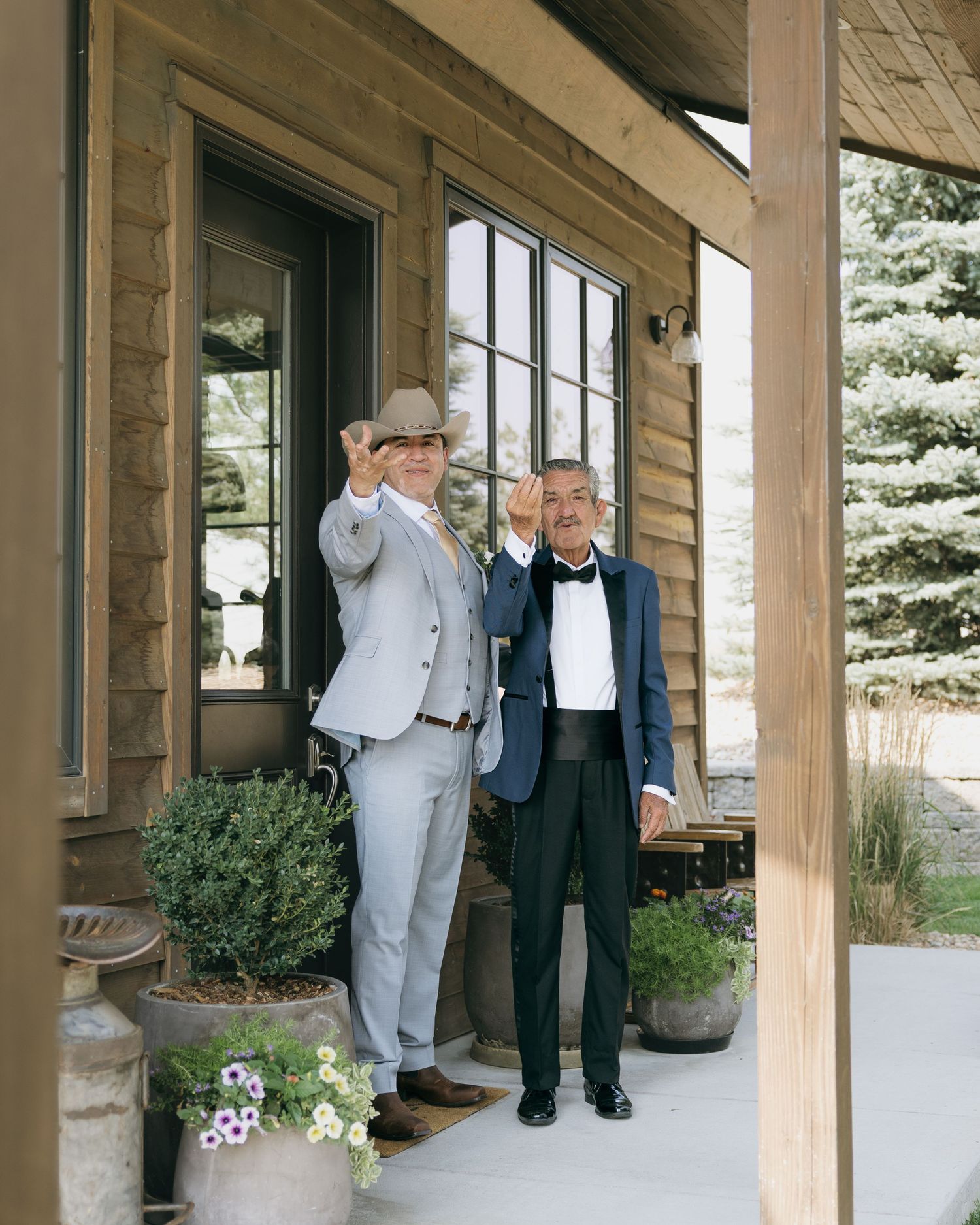 Two men in formal attire tip their hats on a rustic lodge porch, one in a gray suit with cowboy hat.