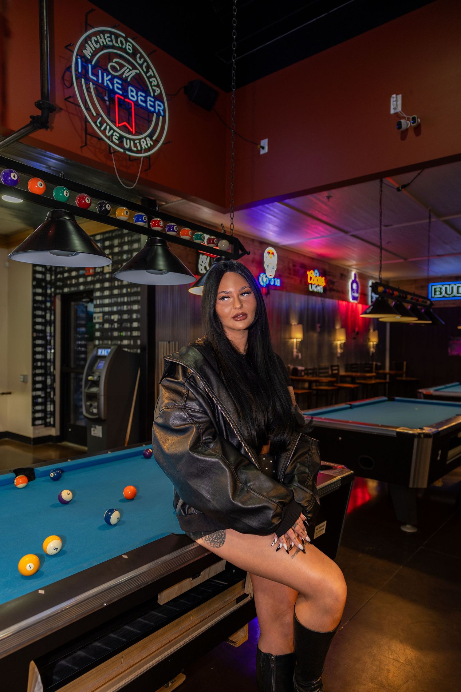 Woman in black leather jacket sitting on pool table in neon-lit bar.