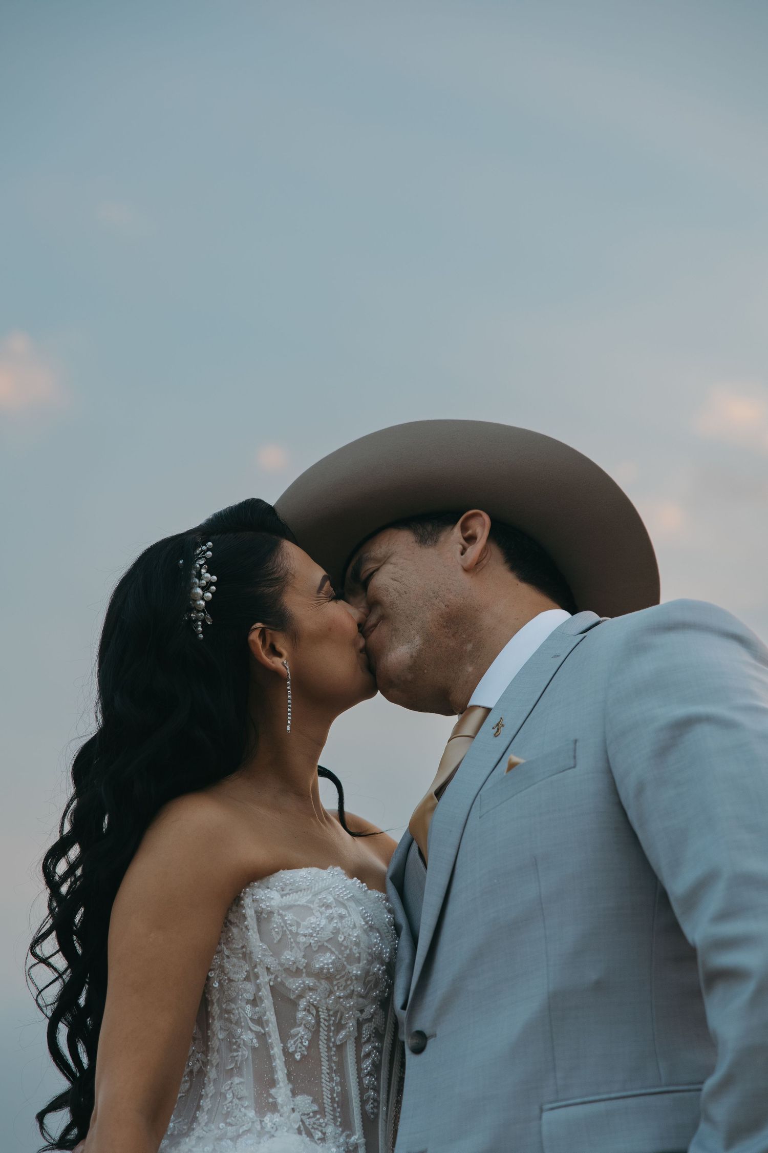 Bride and groom sharing a kiss, groom wearing a tan cowboy hat against a soft blue sky.