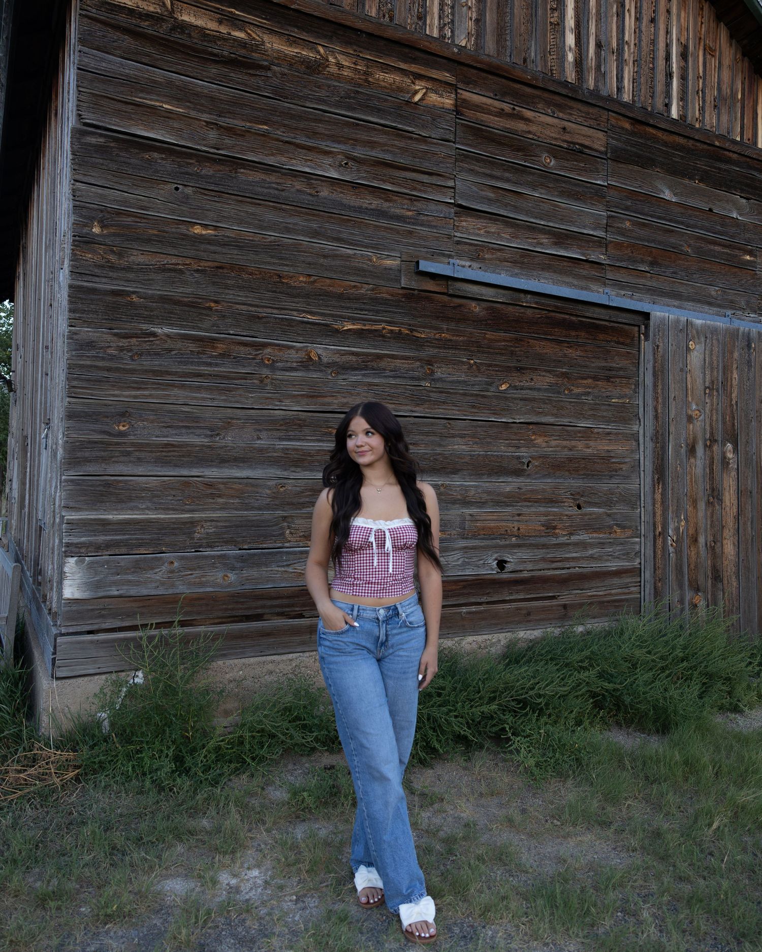 Young woman in pink crop top and flared jeans stands before weathered wooden barn wall. Senior Session