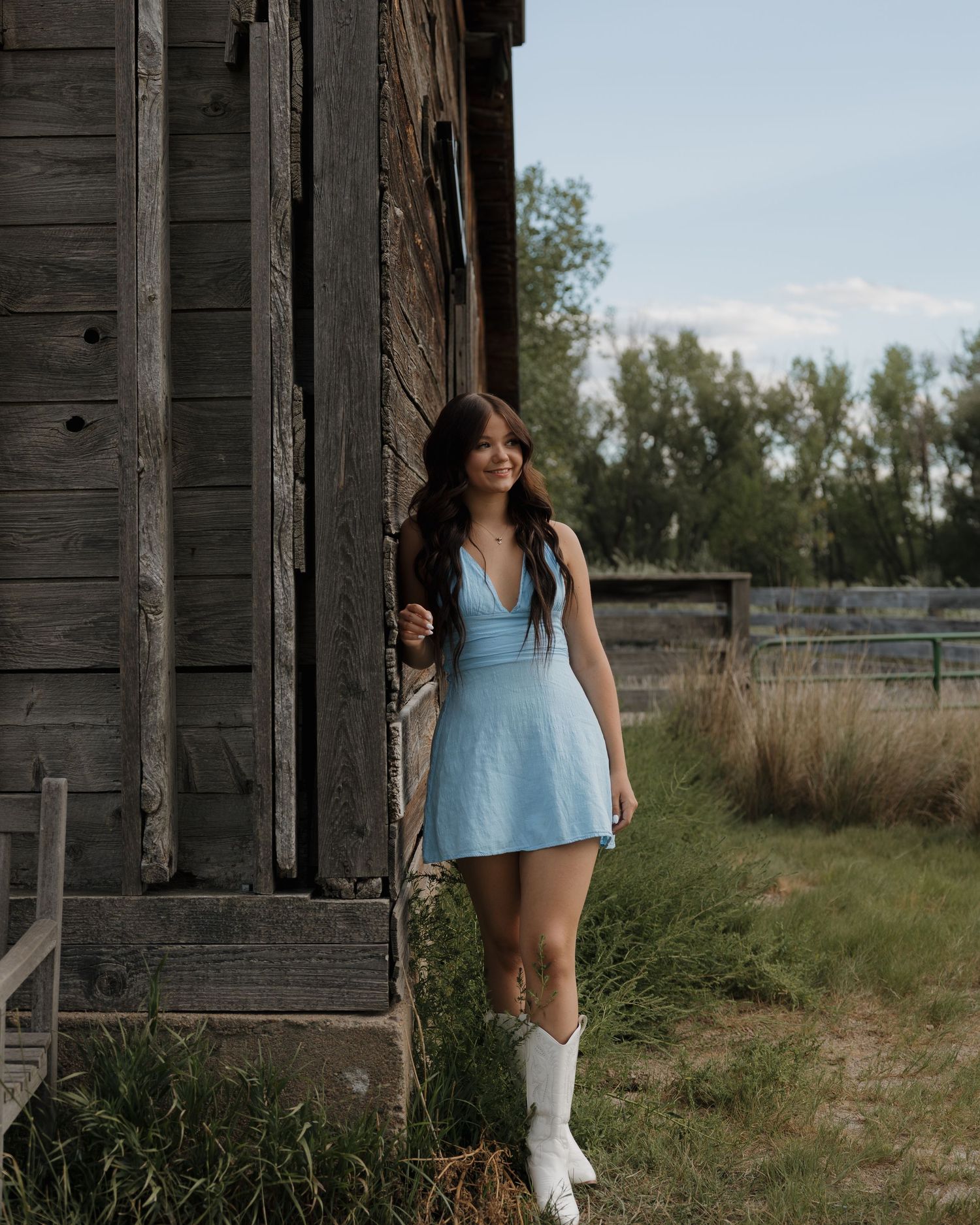 Young woman in light blue mini dress and white cowboy boots poses by rustic wooden barn.