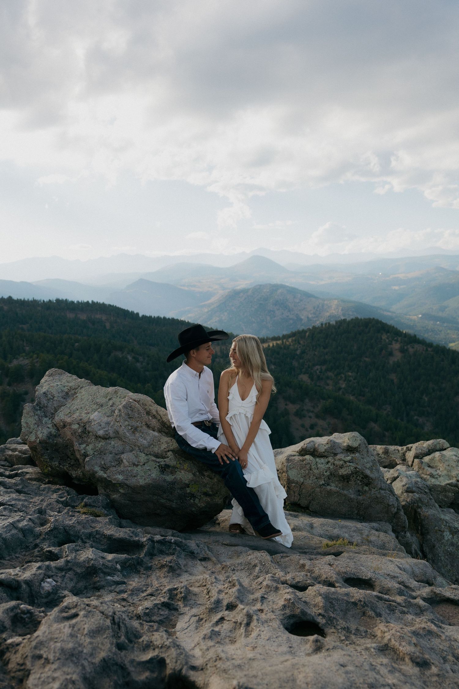 Couple in western attire sits on mountain summit rocks with panoramic forest valley view.