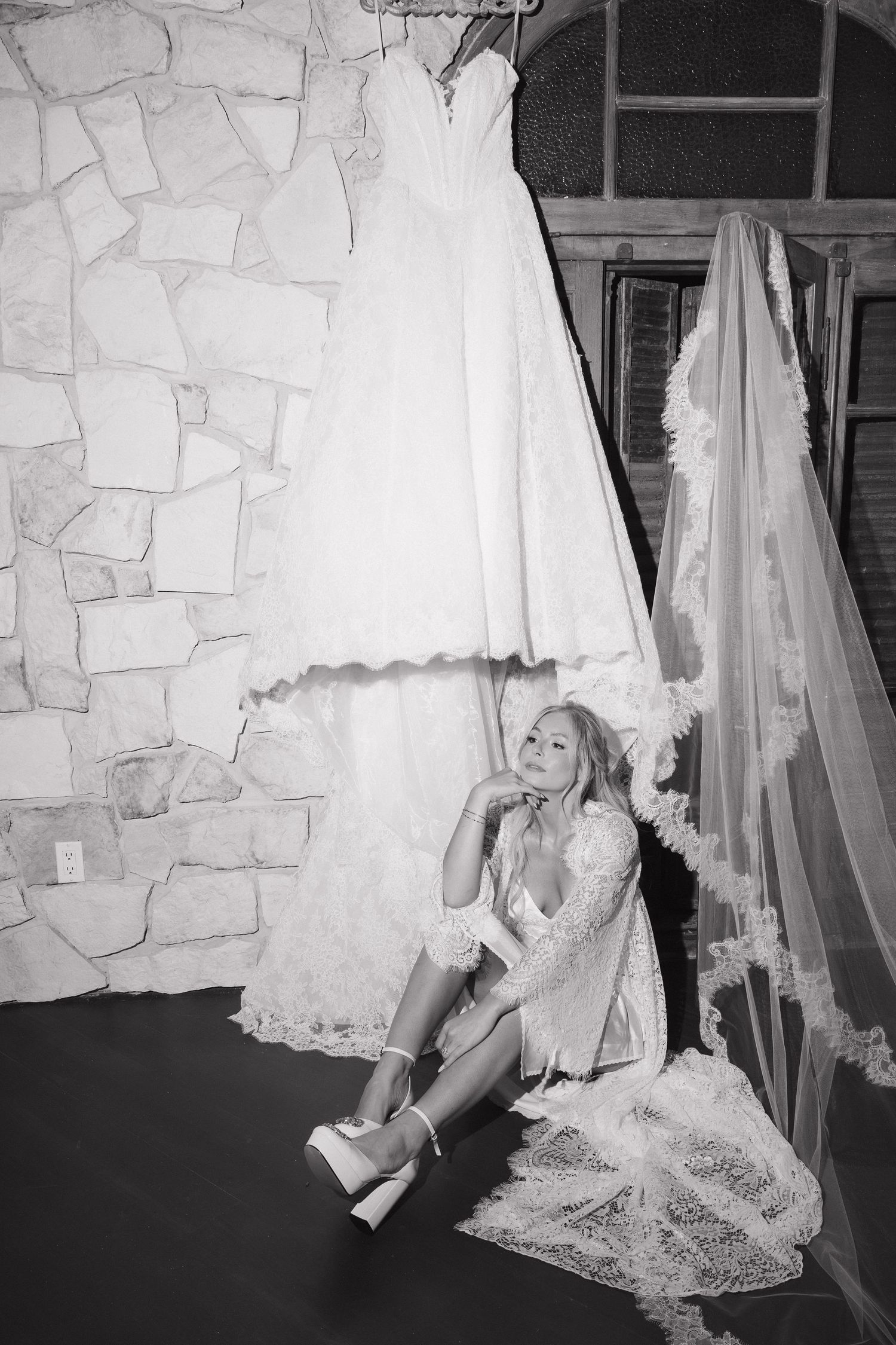Black and white photo of bride sitting on floor beside hanging wedding dress and lace veil.