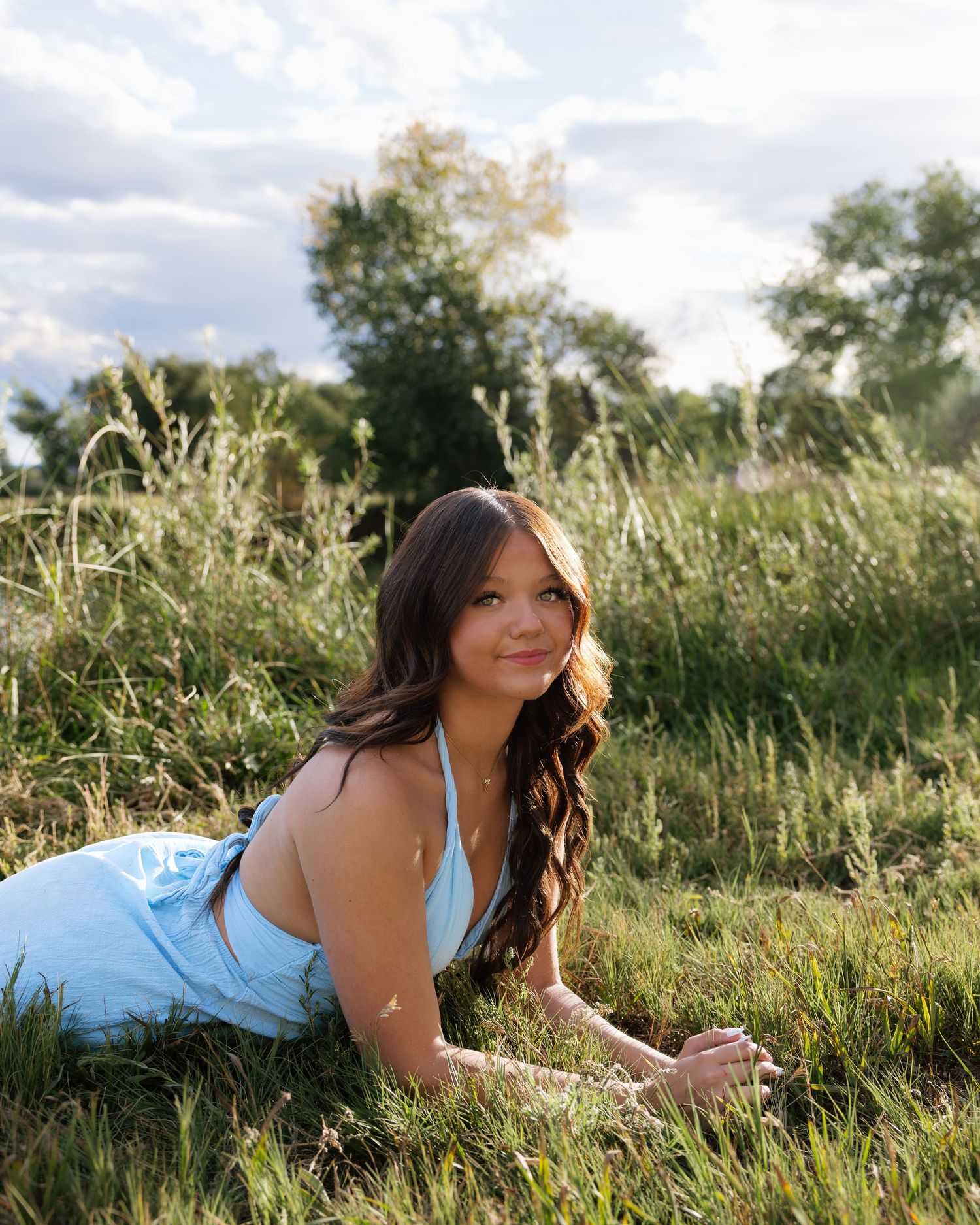 Young woman in light blue dress lies in tall grass field with trees in background.