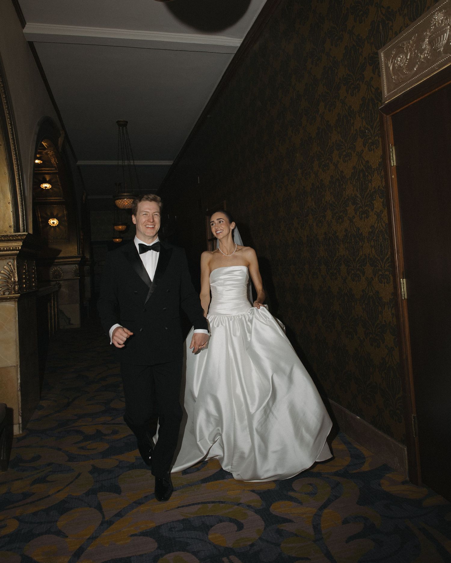 Bride and groom walking hand in hand down ornate hotel corridor in formal wedding attire.