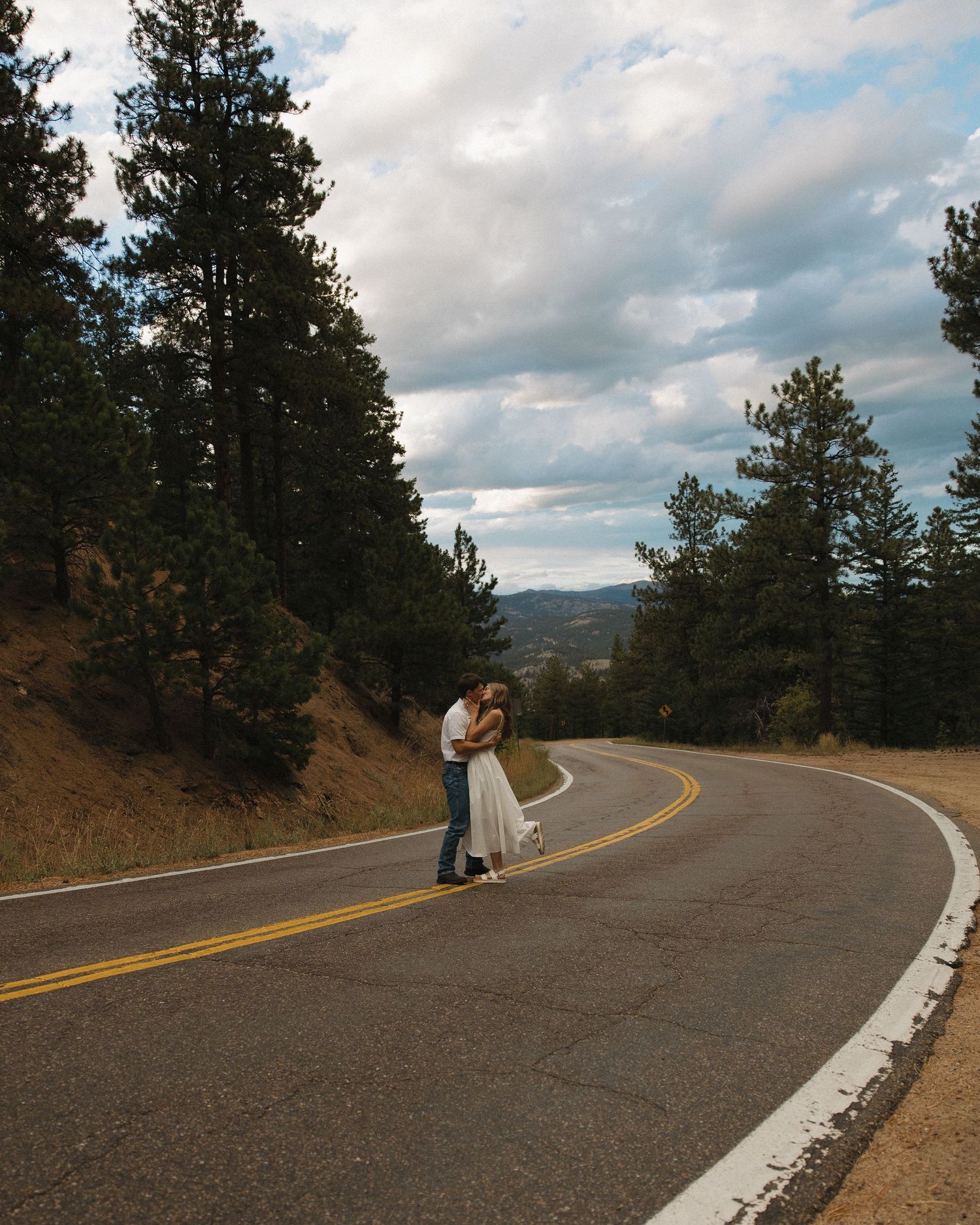 Bride and groom embrace on winding mountain road surrounded by tall pine trees under cloudy sky.