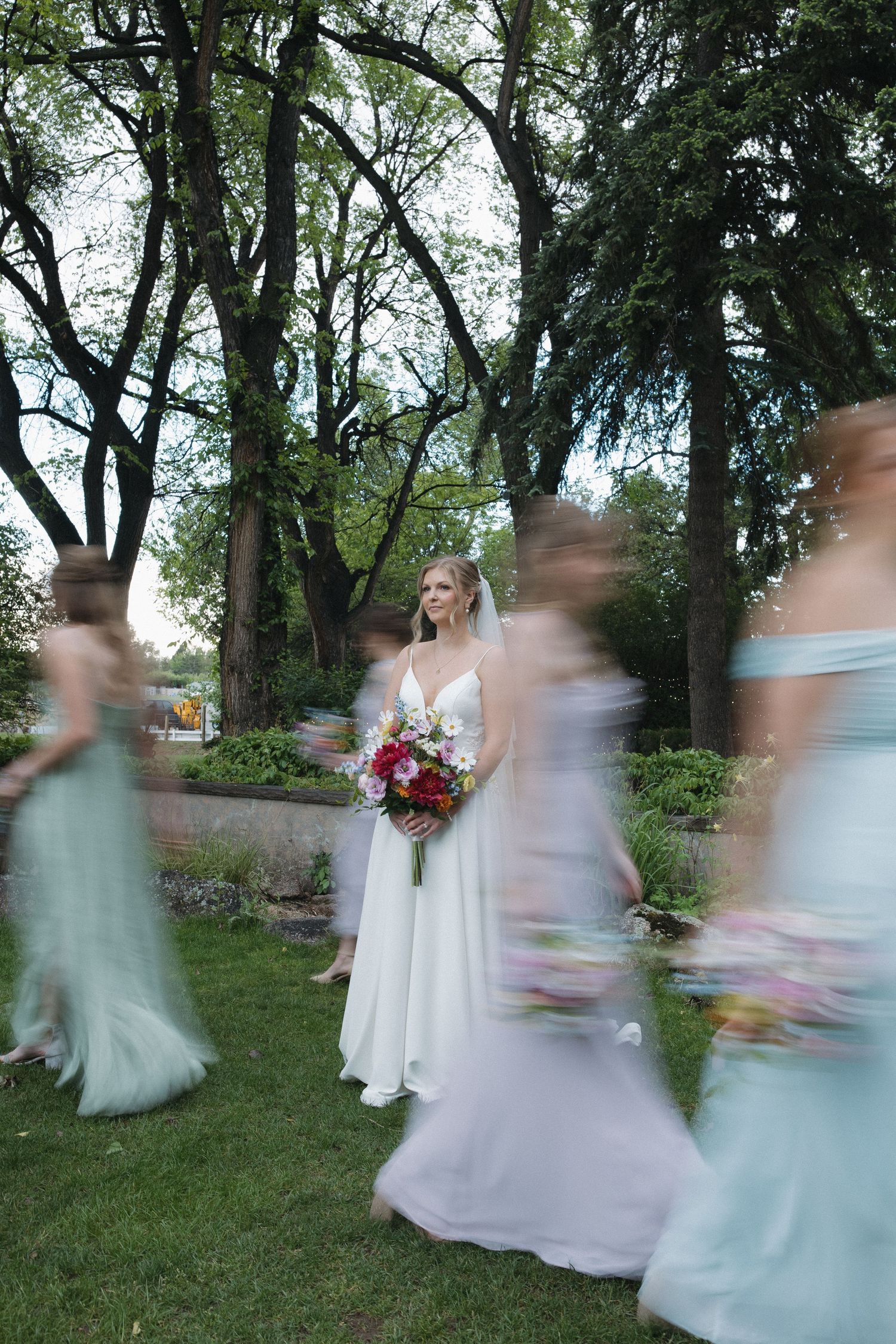 Bride in white gown holds colorful bouquet surrounded by blurred bridesmaids in sage green dresses in a lush garden.