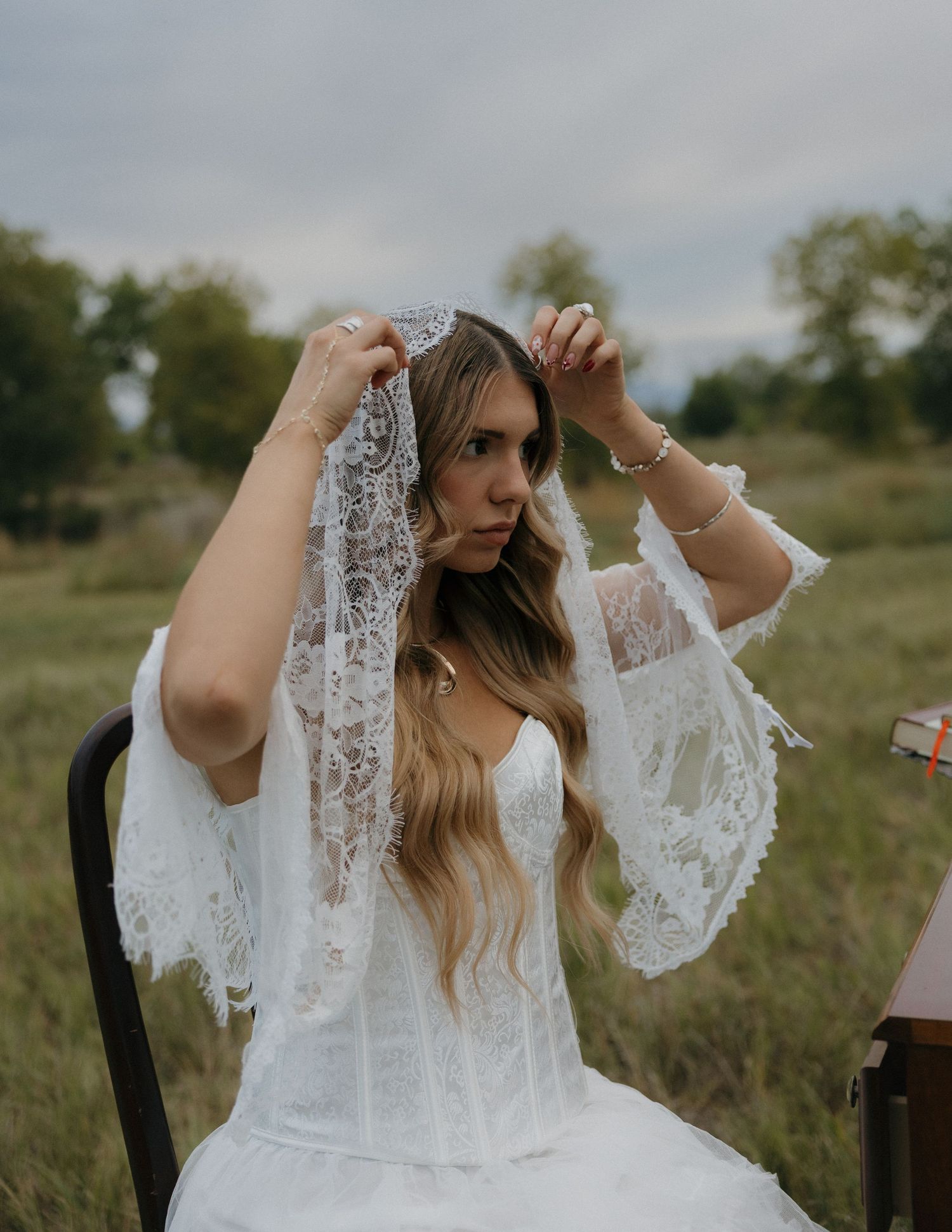 Bride placing lace veil over head wearing white bridal gown in an outdoor field setting.