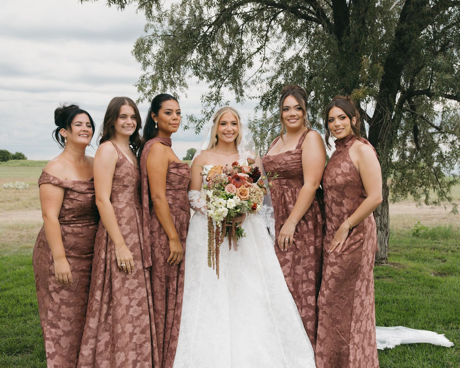 Bride in white gown poses with five bridesmaids in dusty rose floral dresses outdoors.