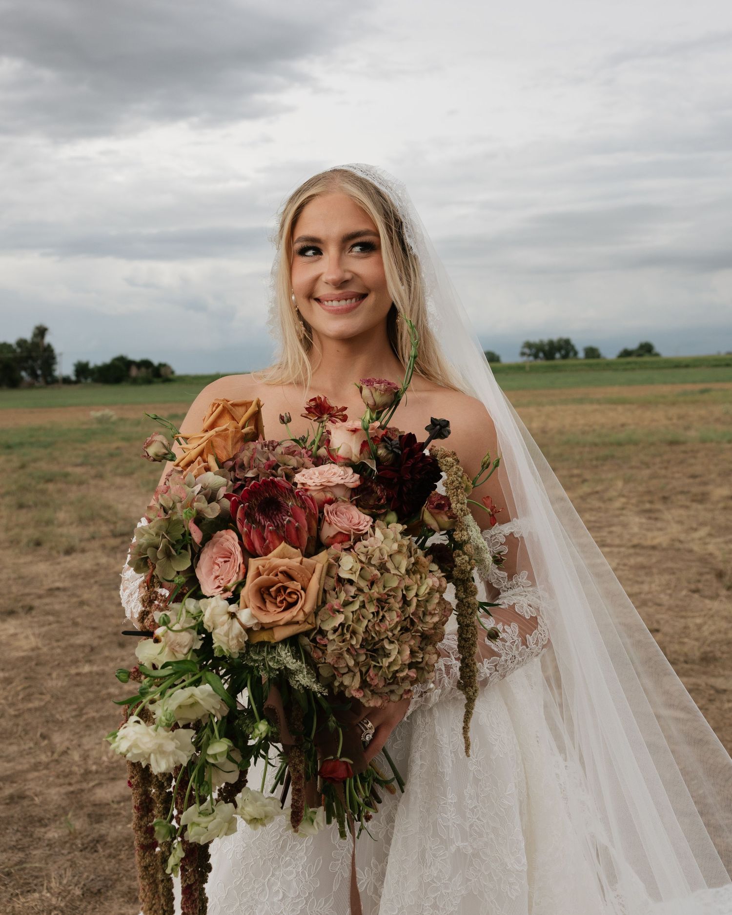 Smiling bride in lace wedding dress holds large bouquet of roses and hydrangeas in an outdoor field setting.