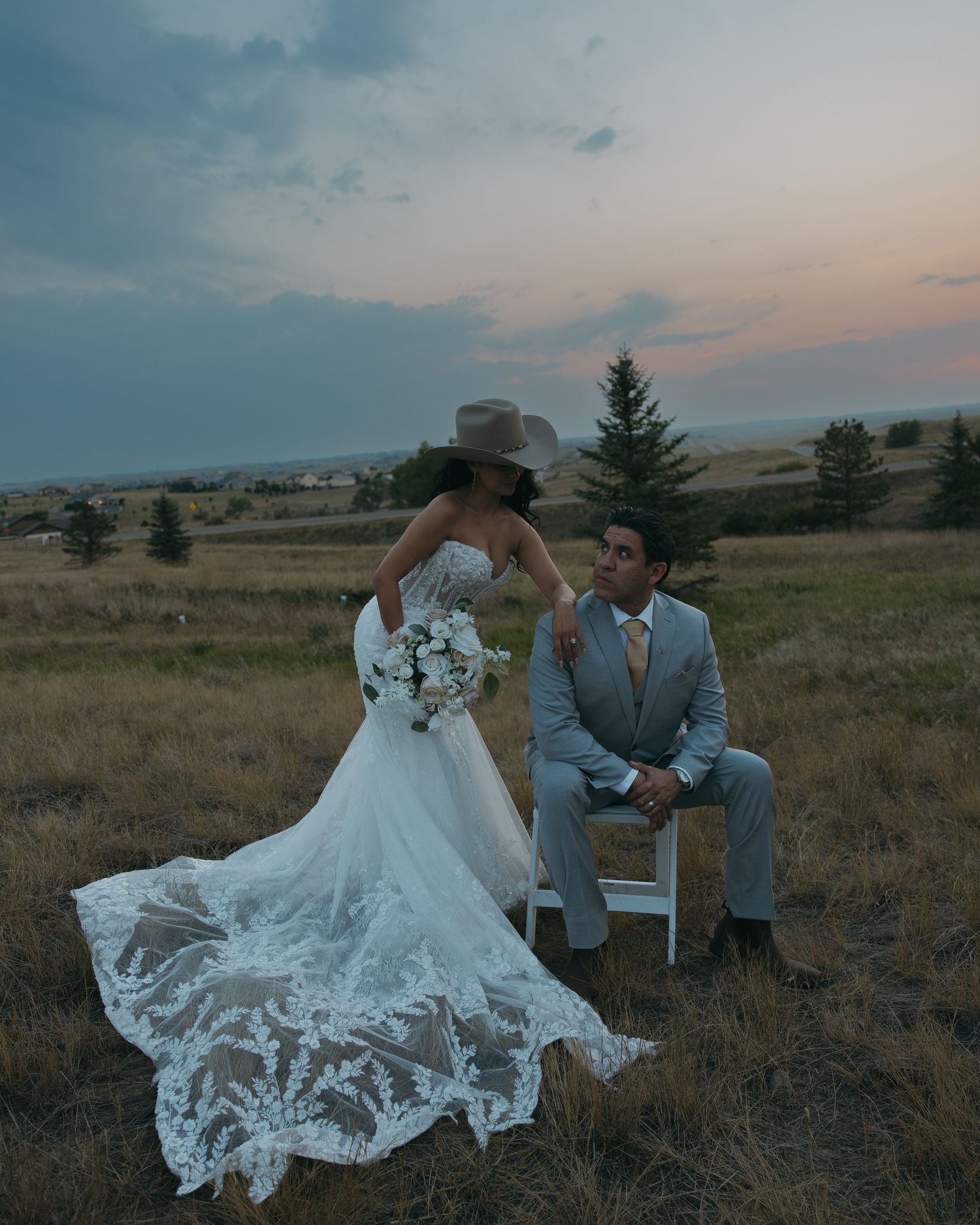 Bride in lace gown and cowboy hat stands beside seated groom at sunset on open plains.