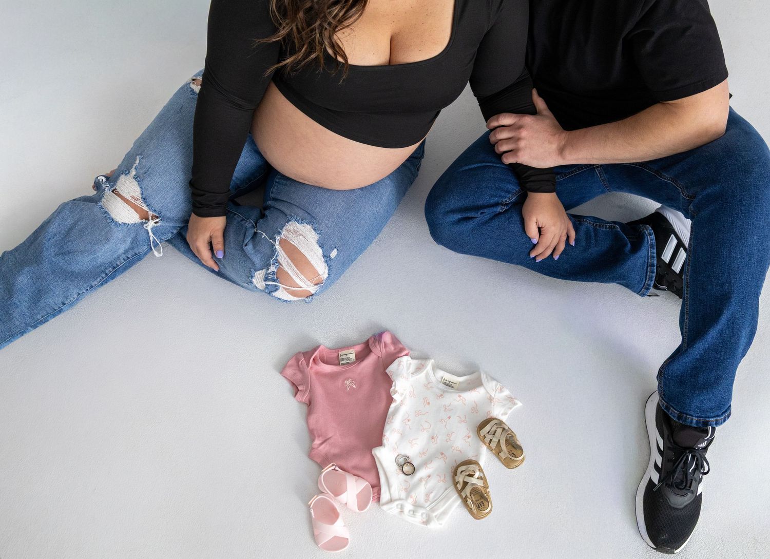 Expecting couple sitting on floor with baby clothes and shoes between them.