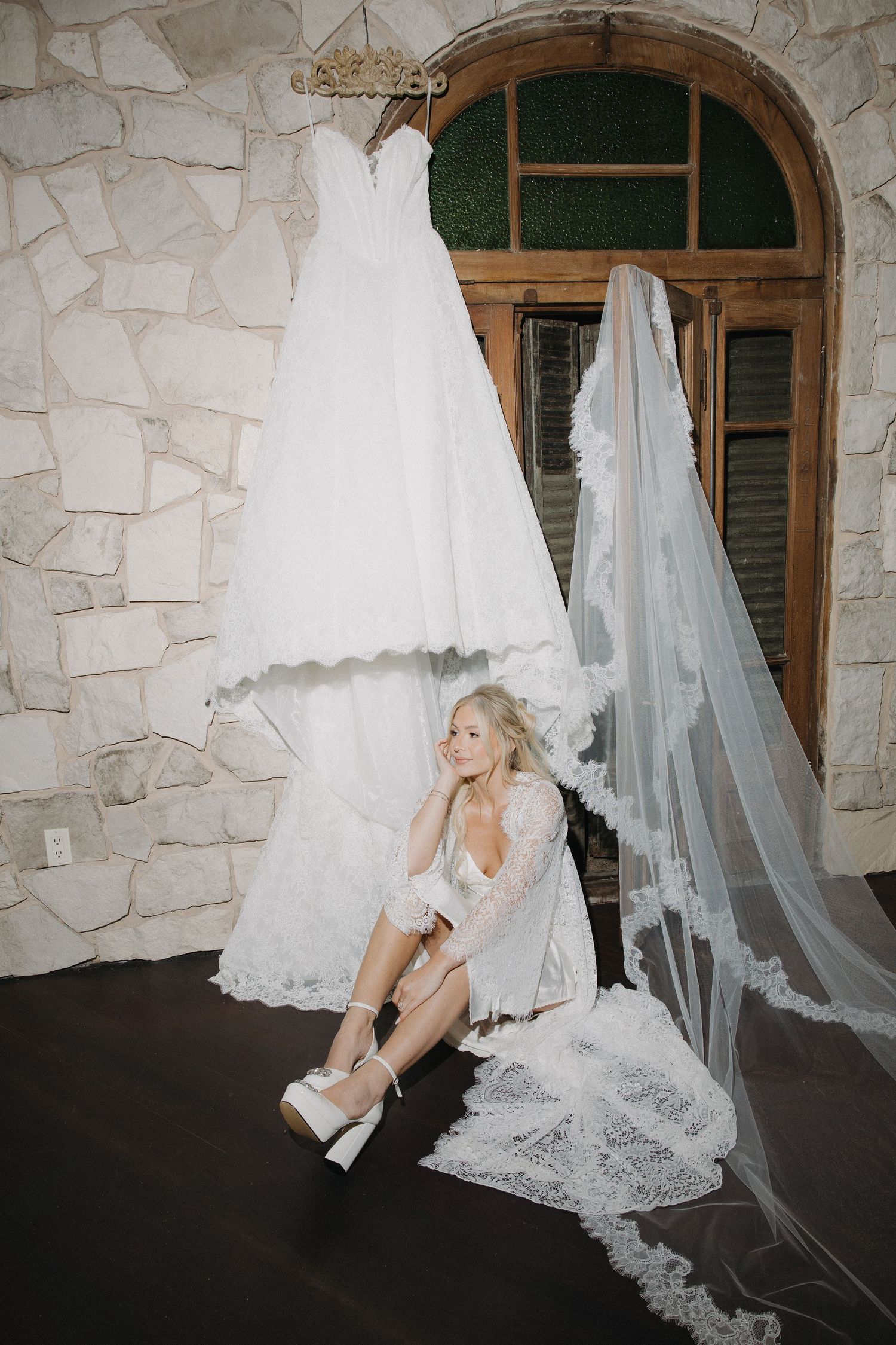 Blonde bride in lace robe sits on floor putting on heels beside hanging wedding gown and veil.
