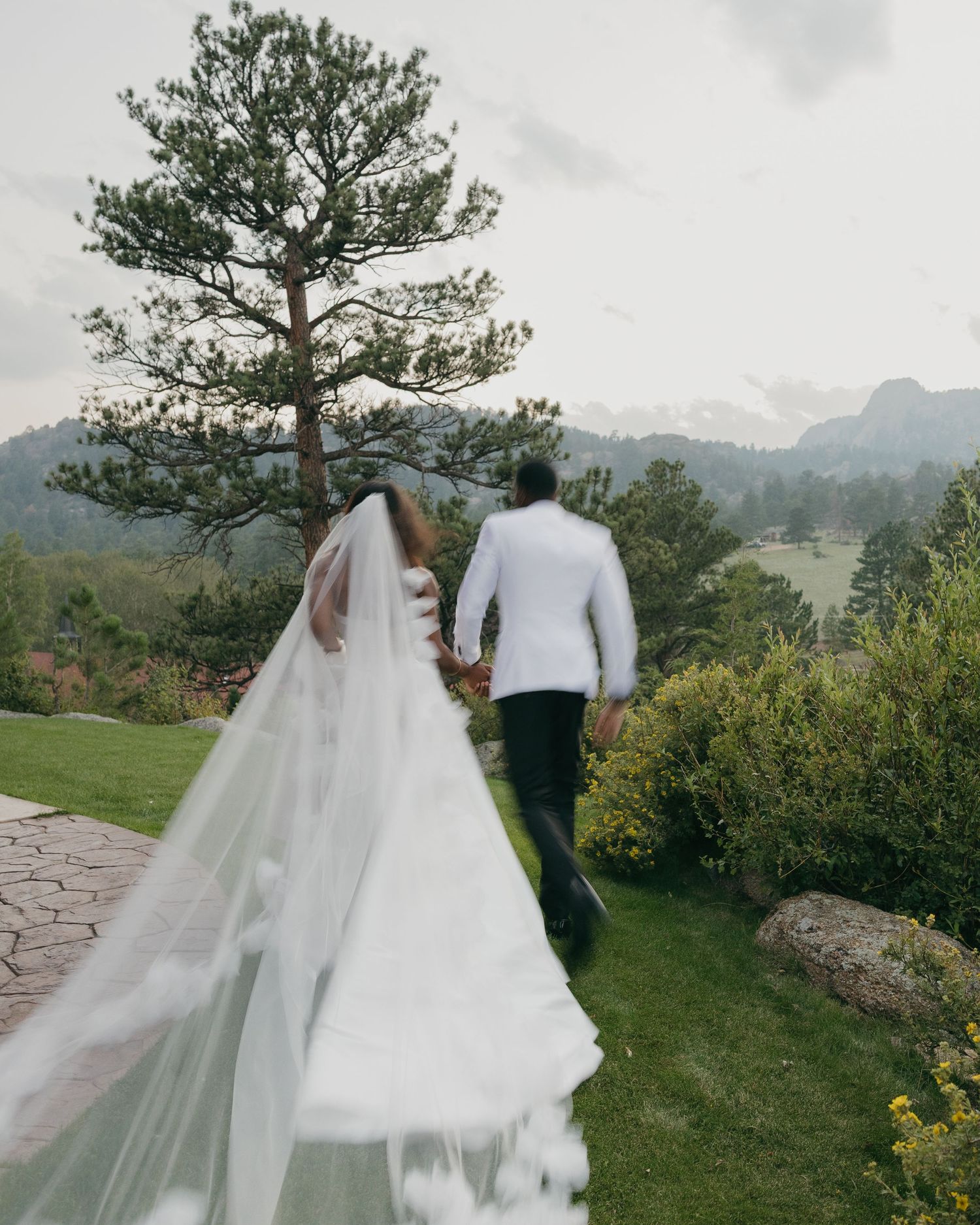 Bride and groom walking hand in hand through scenic mountain garden with flowing veil.