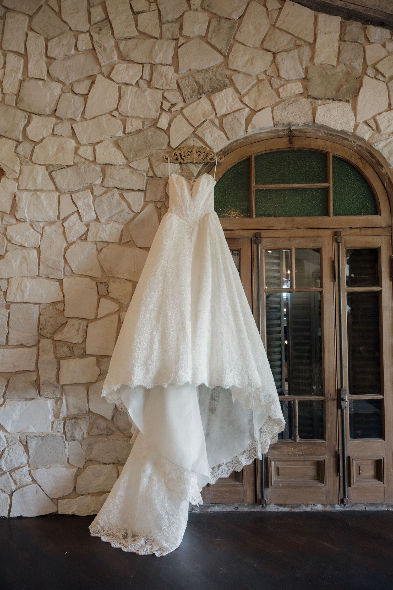 Elegant lace ball gown wedding dress hanging on stone wall near arched wooden doors.