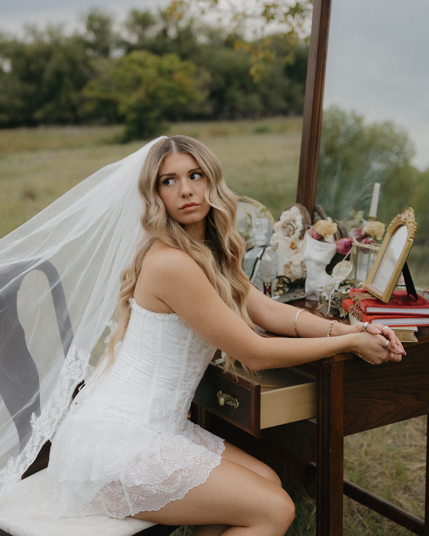 Bride in white dress and veil sits at vintage vanity desk in open grassy field.