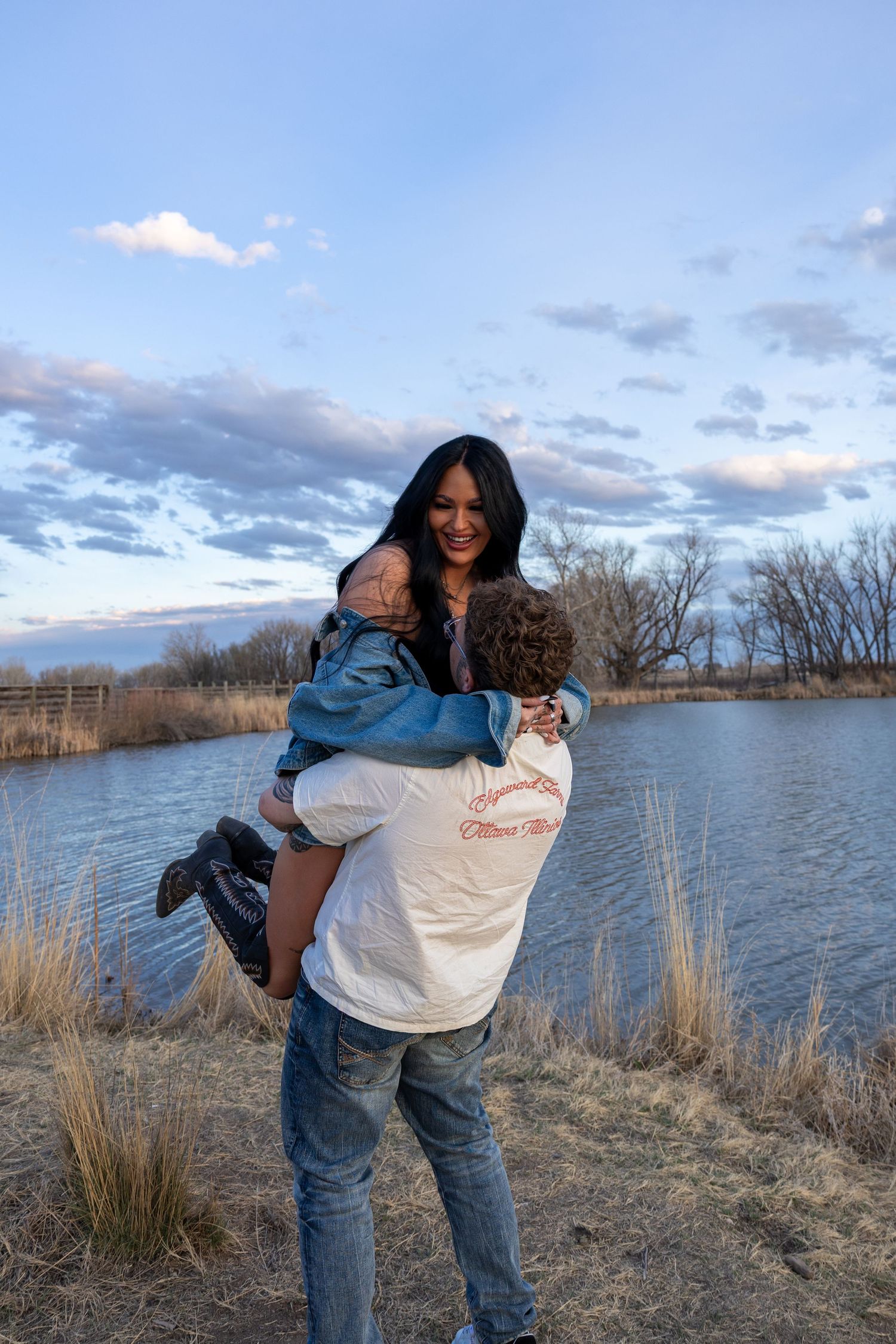 Man lifting woman near pond at sunset in rustic outdoor setting.