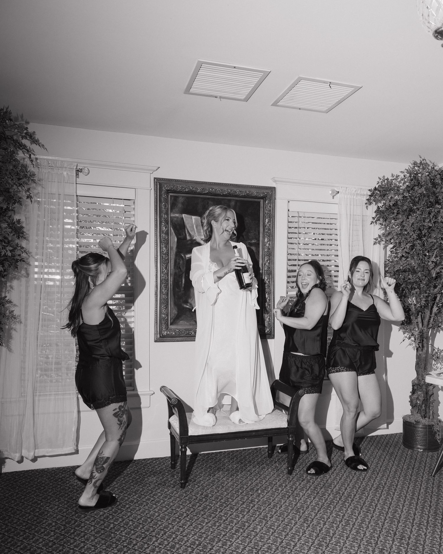 Black and white photo of bride celebrating with bridesmaids, holding champagne bottle in white robe.