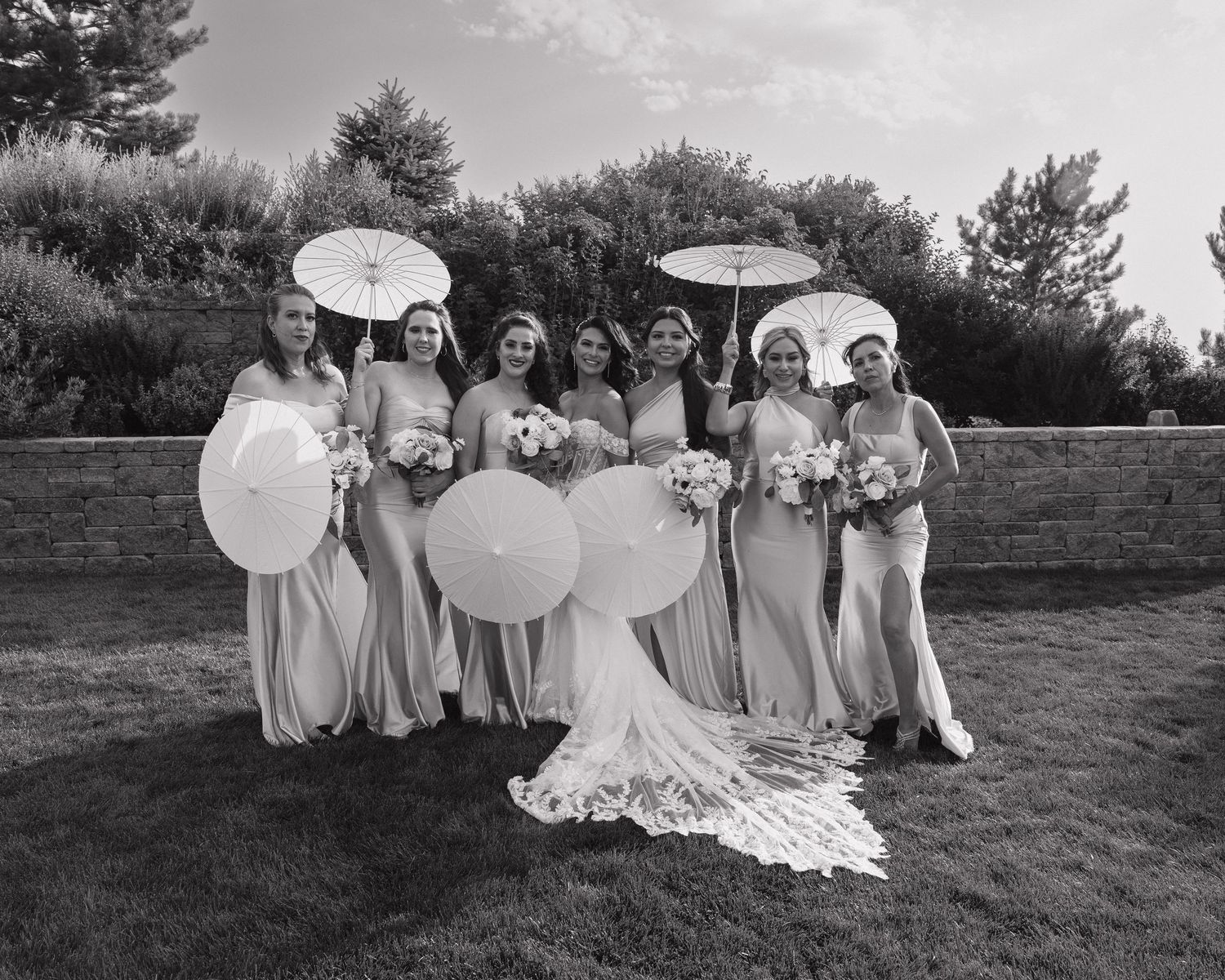 Bridesmaids in matching gowns hold parasols and bouquets for elegant outdoor wedding party portrait.