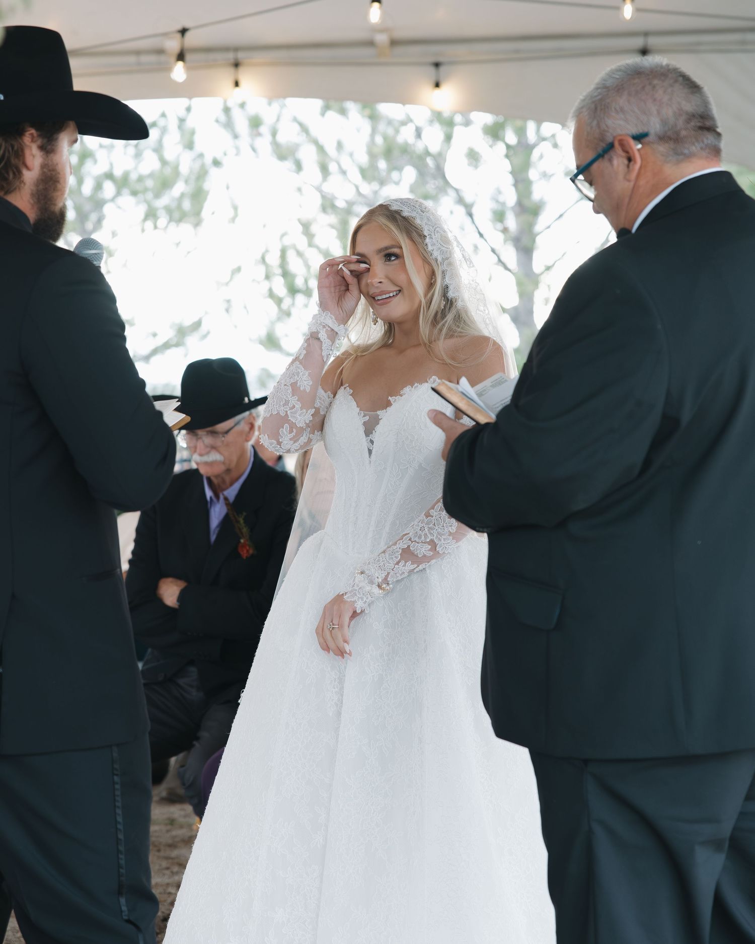 Emotional bride wiping tears during outdoor wedding ceremony wearing long-sleeve lace gown.