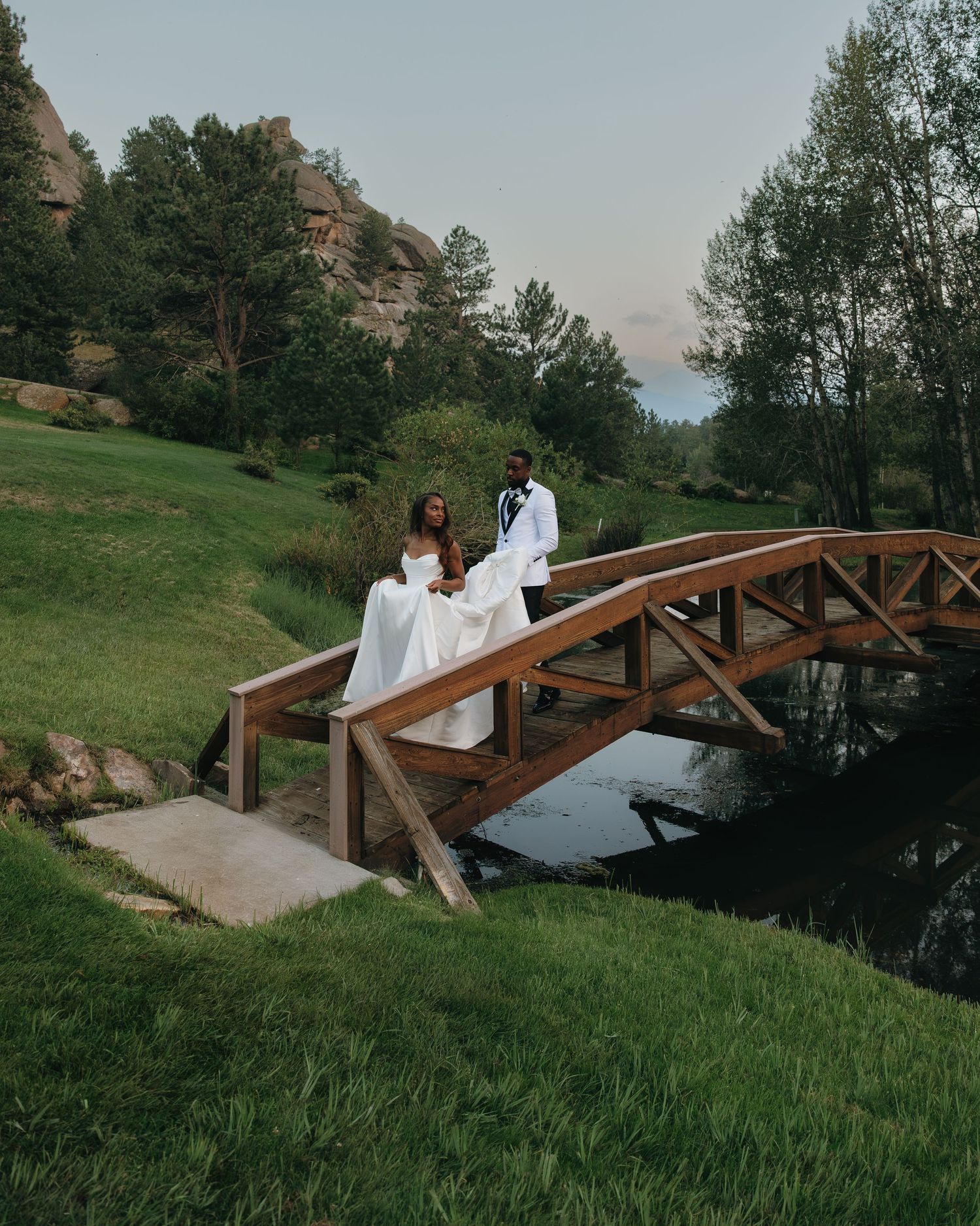 Wedding couple in white attire poses on a rustic wooden bridge surrounded by lush greenery.