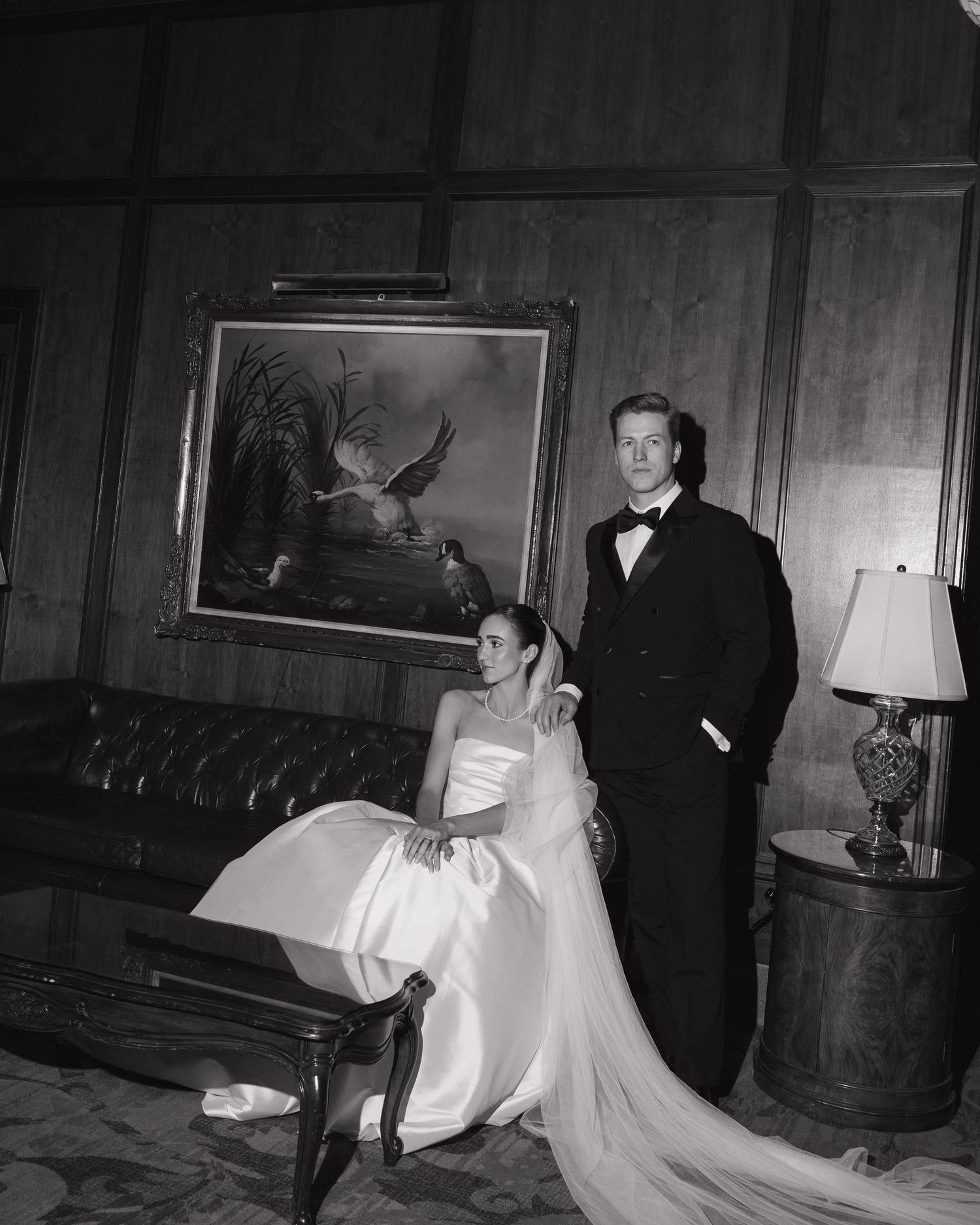 Black and white portrait of bride in strapless ballgown and groom in tuxedo in wood-paneled room.